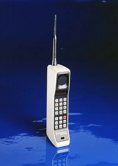 A vintage mobile phone with an extendable antenna standing upright on a reflective blue surface.