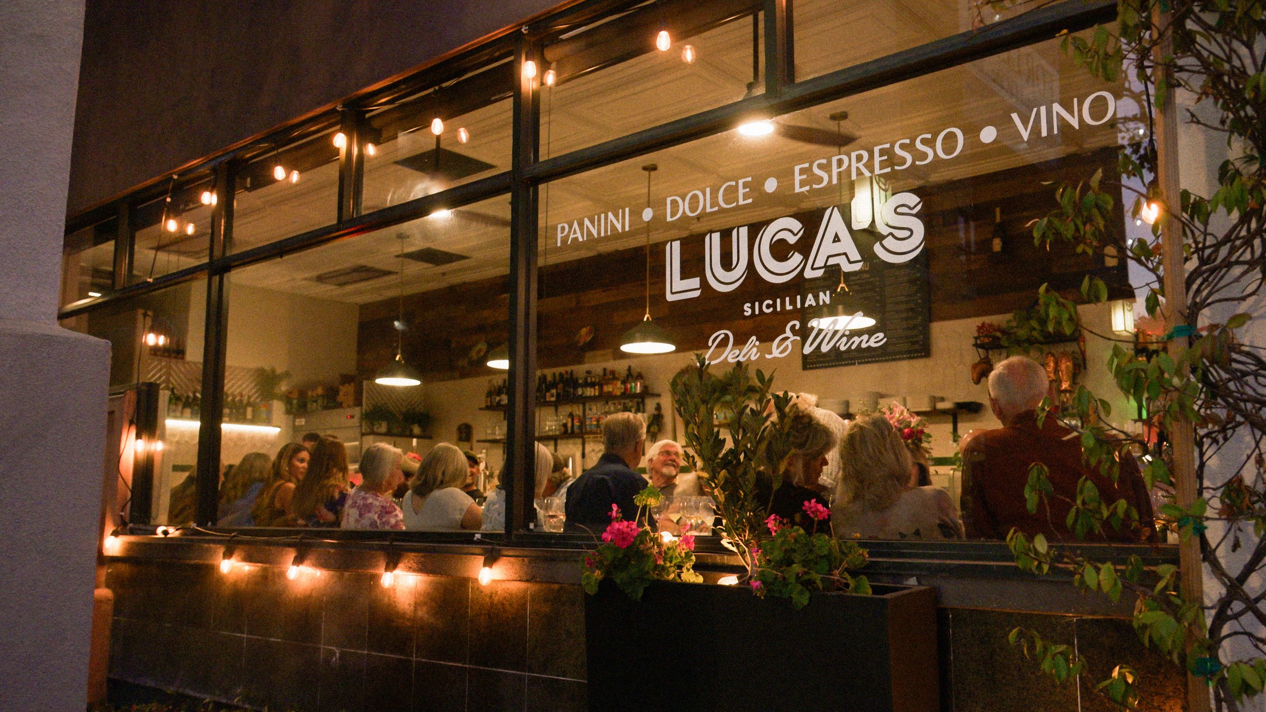 View through a restaurant window showing a group of people dining inside, with decorative lighting, shelves of bottles, and sign with menu items including panini, espresso, wine, and desserts.