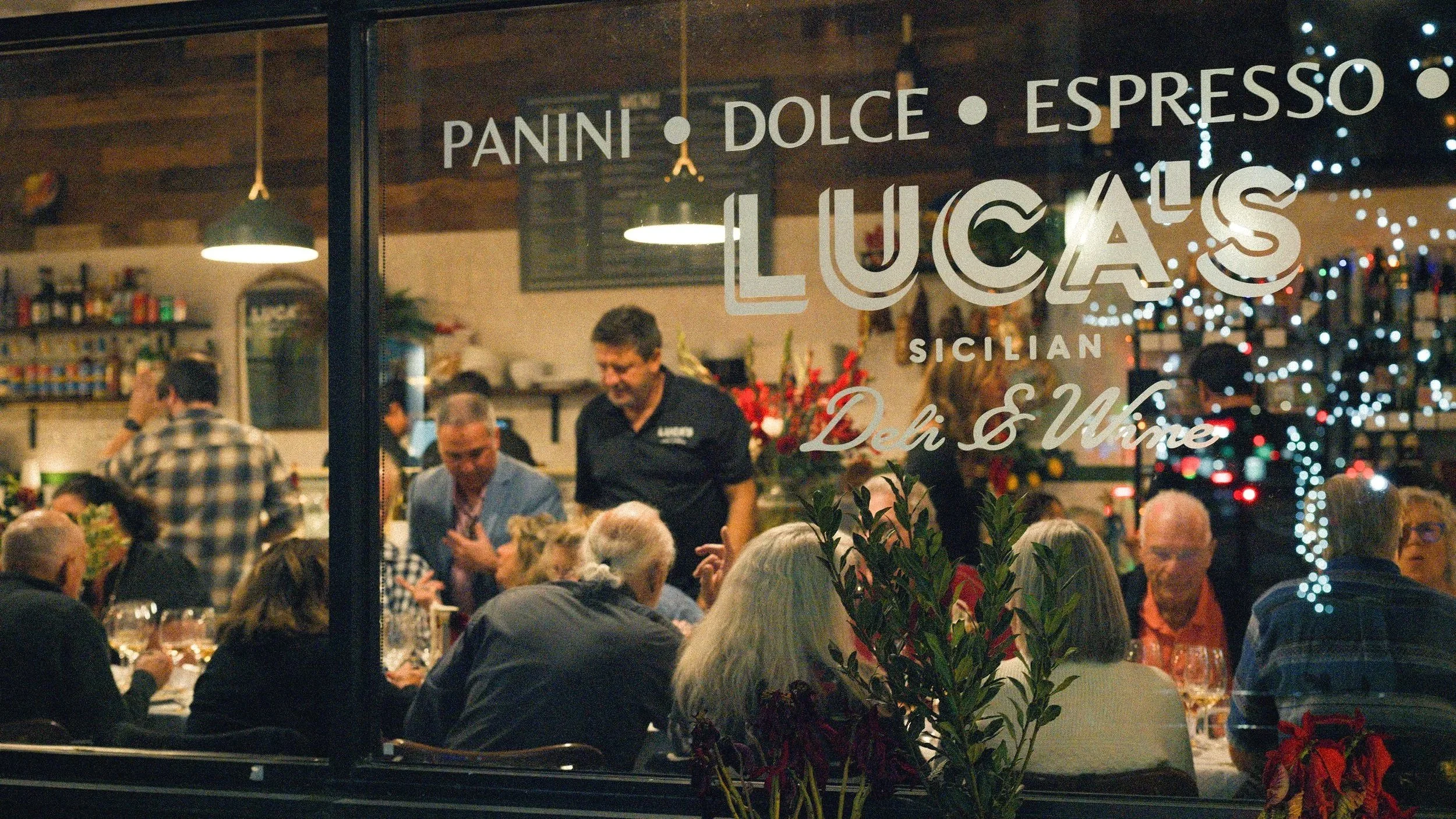 A busy restaurant scene seen through a window with signs advertising panini, dolce, espresso, and wine. Inside, diners are seated at tables, and staff are serving food. Christmas decorations and string lights are visible, indicating a festive atmosphere.