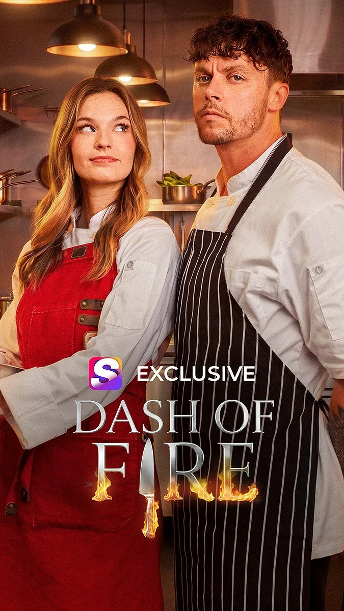 Two chefs, a woman with long red hair and a man with curly brown hair, standing in a professional kitchen. The woman is wearing a white chef's coat and a red apron, while the man is wearing a white chef's coat with a black striped apron. The background shows kitchen equipment and warm lighting. The text on the image reads 'Exclusive Dash of Fire' with flames incorporated into the design.