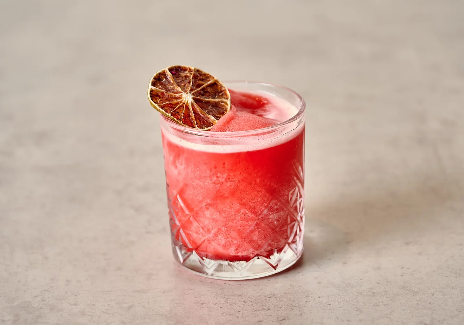 Red cocktail in a short glass with a dried citrus garnish.