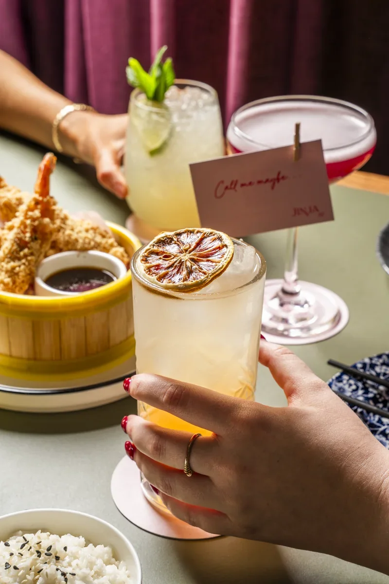 Three cocktails held above a dining table in front of Jinja’s pink curtains.