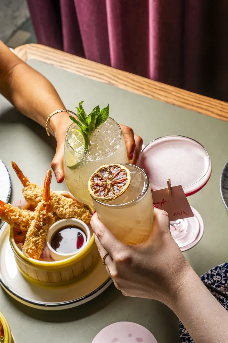 Two cocktails held together above a table of sharing dishes.