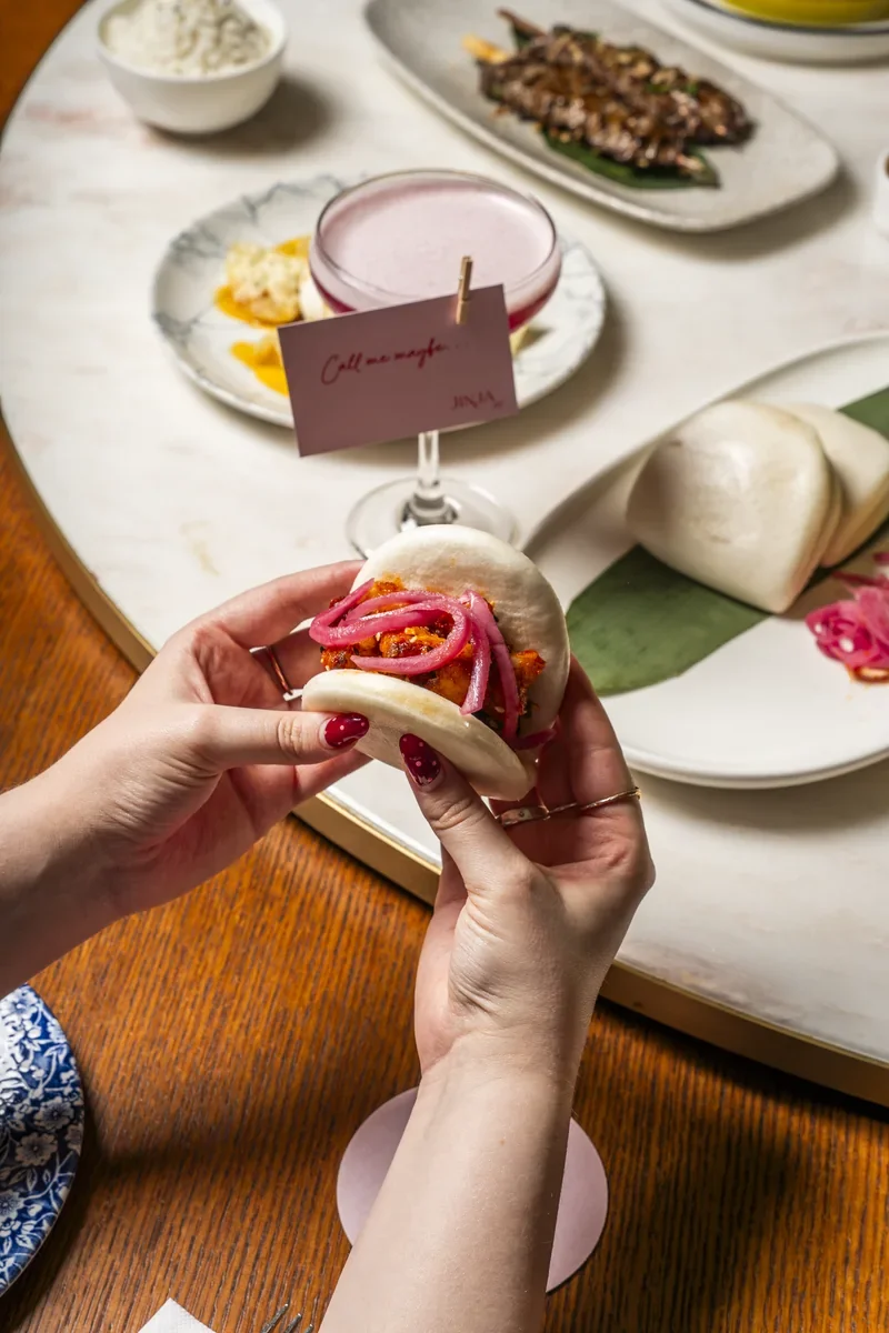 Hands lifting a filled bao bun above a table of sharing dishes.