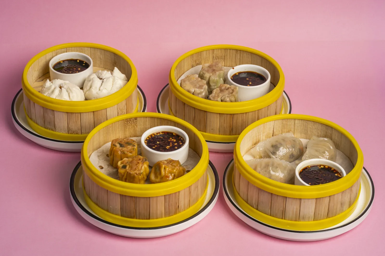 Dumplings and buns in four bamboo steamer baskets on a pink background.