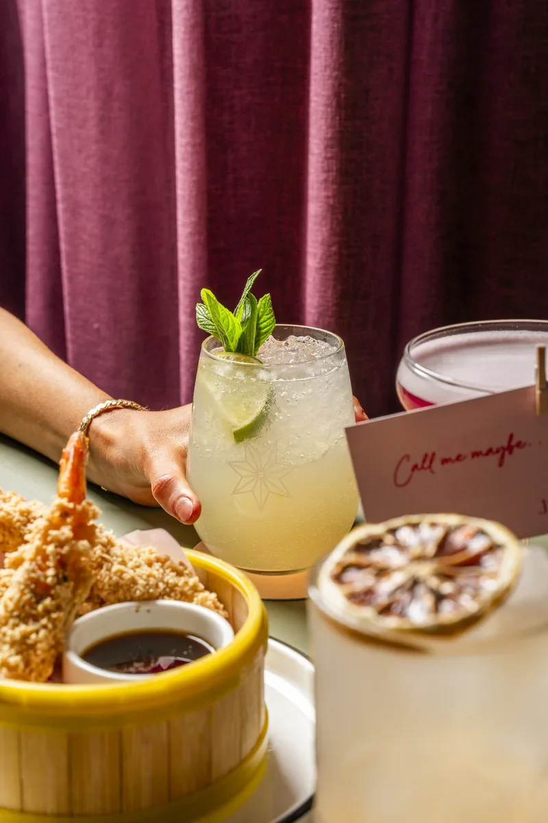 Pale cocktail with a green garnish and a pink stemmed cocktail beside sharing dishes.