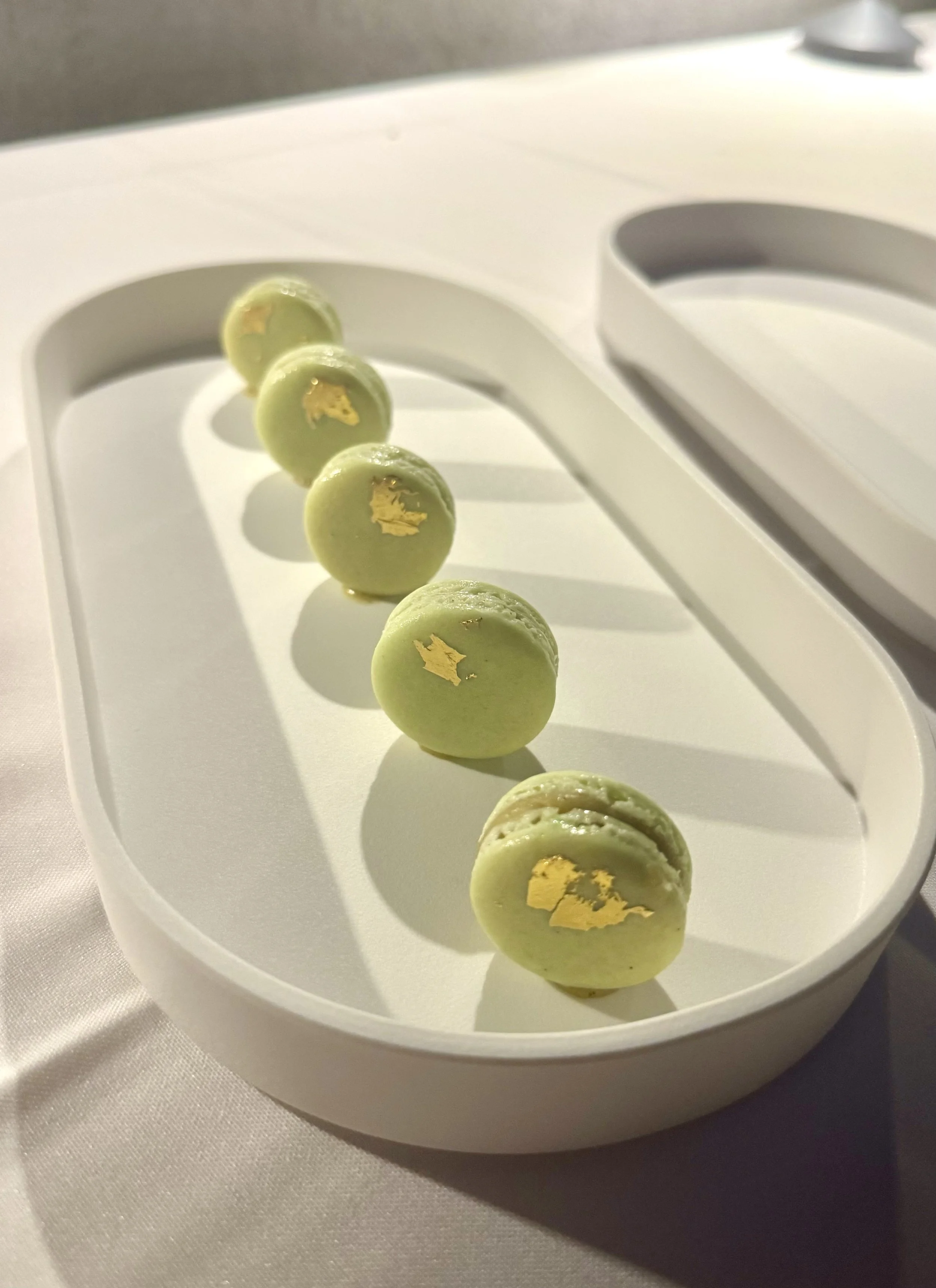 Six green macarons with gold leaf on a white platter, arranged in a line.