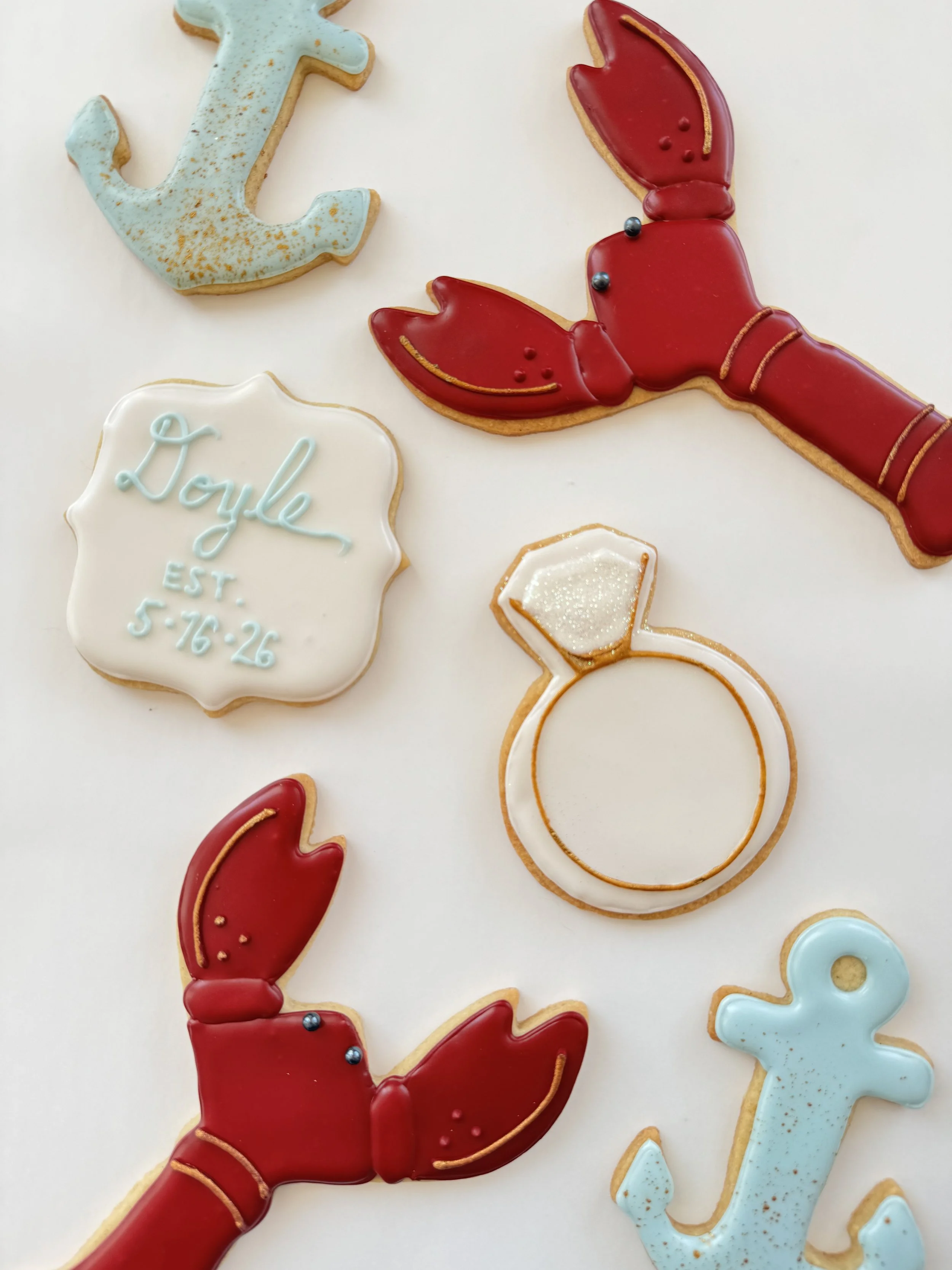 Decorative cookies themed around engagement and the sea, including an anchor, a lobster, a diamond ring, and a personalized cookie with the name 'Doyle' and a date.