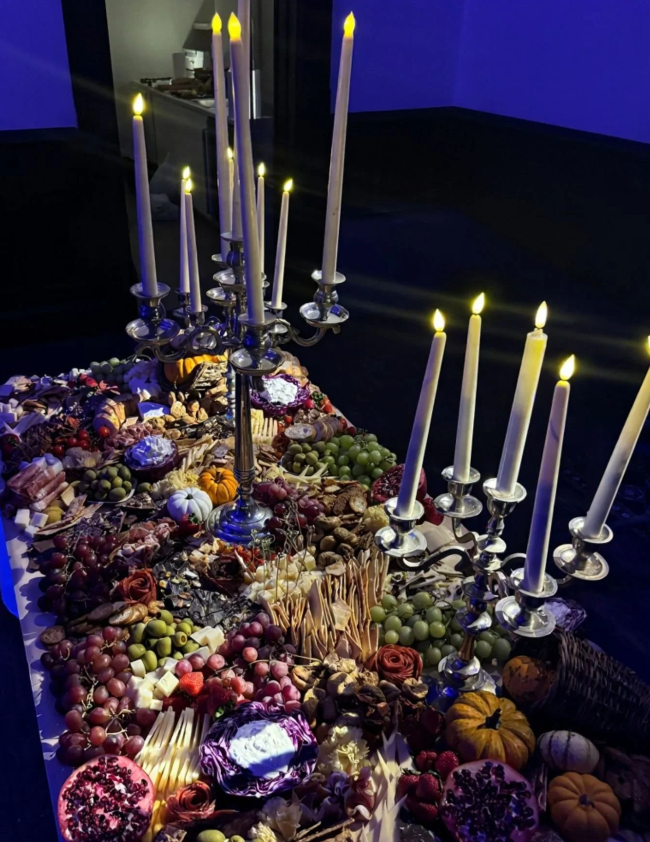 A large table covered with an assortment of food and surrounded by tall candleholders with lit candles, set in a dark room with blue lighting