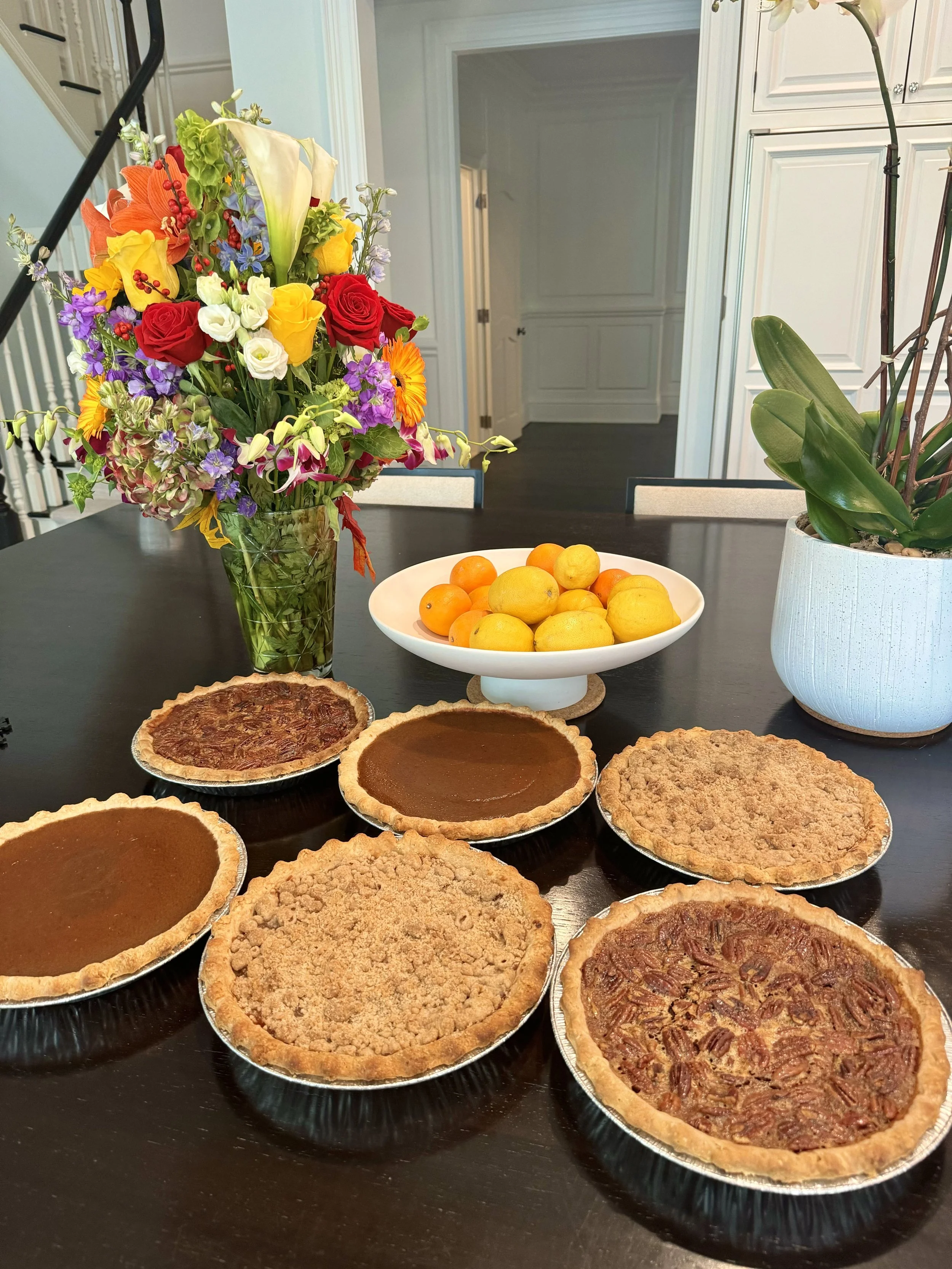 A table with five pies, a bowl of lemons and oranges, a bouquet of colorful flowers, and a potted plant.