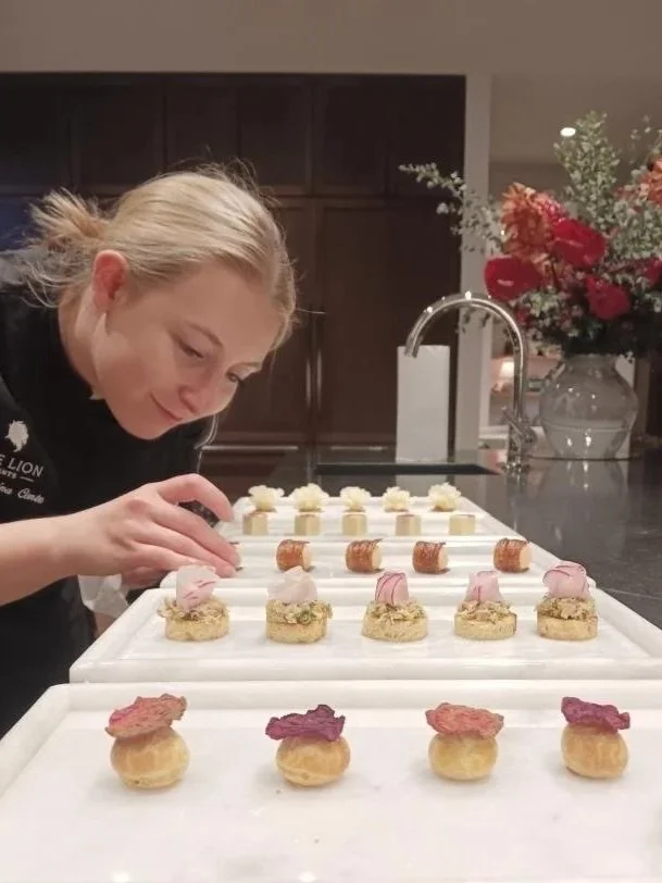 A woman decorating small, elegant pastries on a white tray in a kitchen with dark cabinets and a vase of red and pink flowers in the background.
