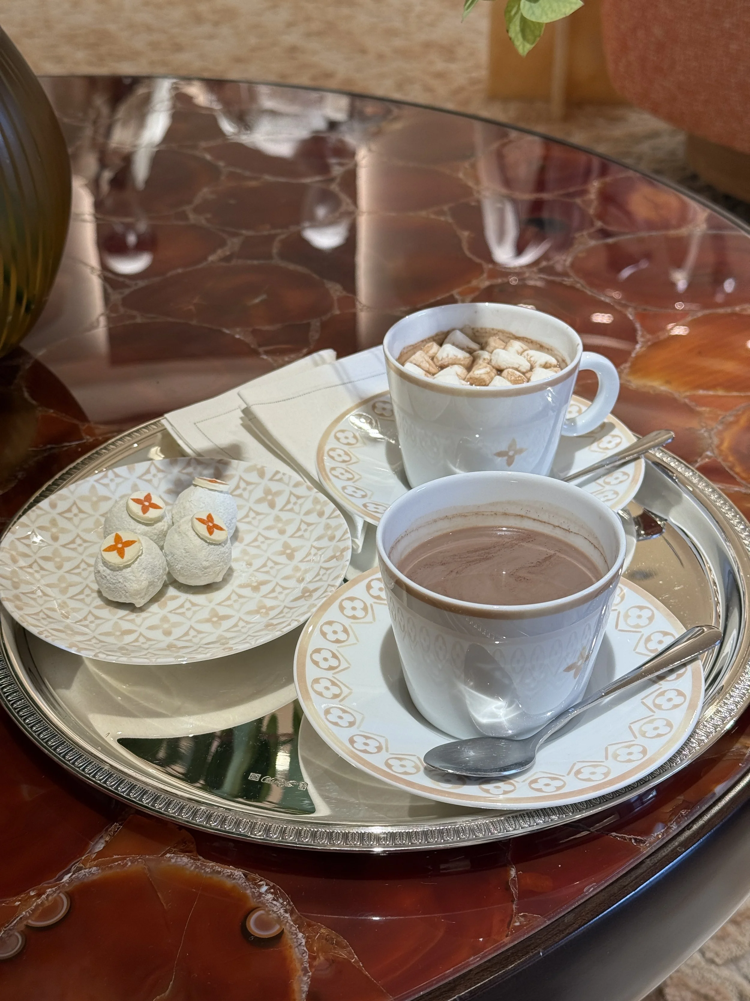 Tray with two cups of hot chocolate topped with marshmallows, a plate of three white chocolates decorated with orange flowers, and a spoon, on a polished marble table.