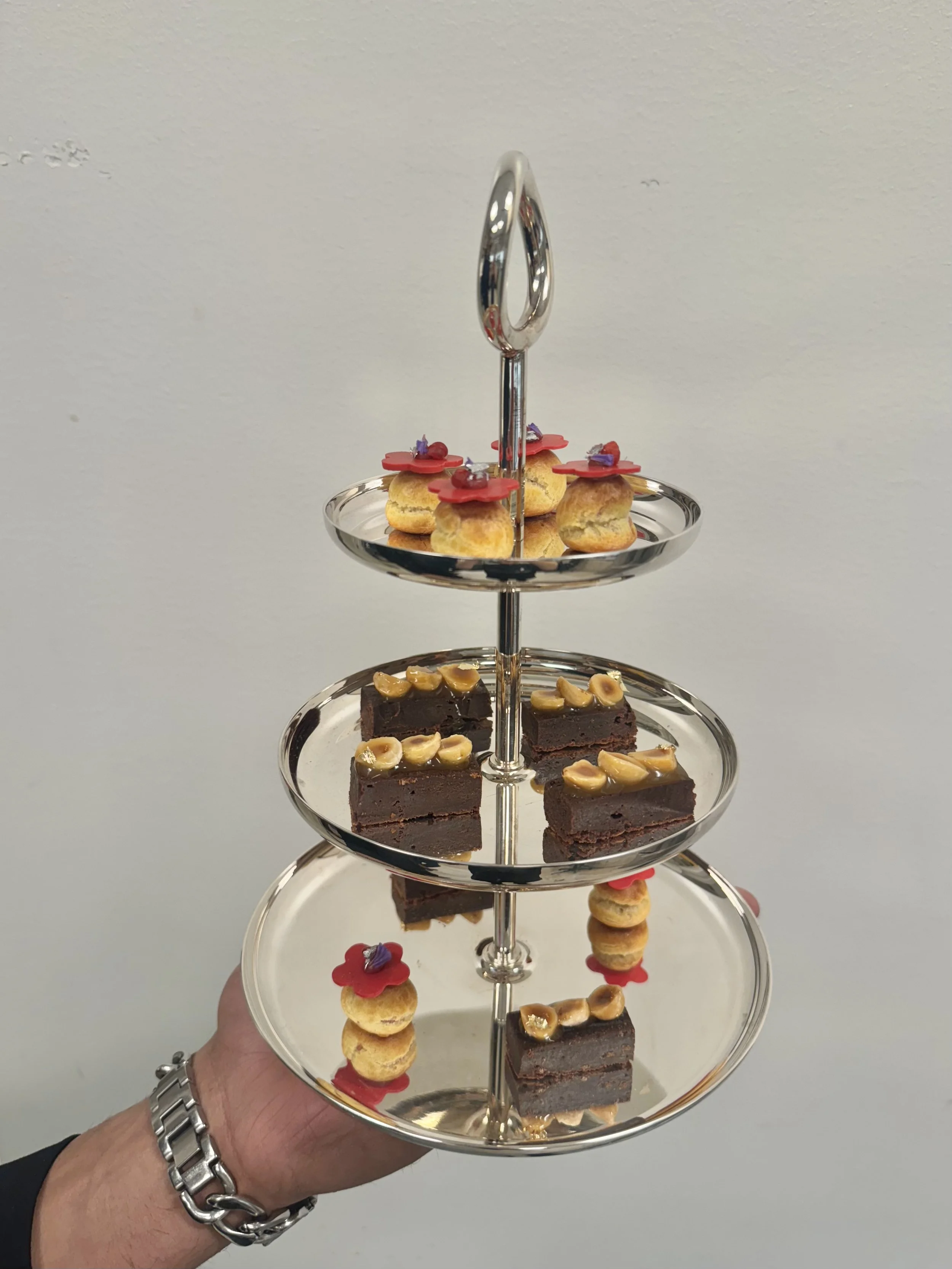 A three-tiered silver dessert tray holds assorted small cakes and pastries, including chocolate brownies with nuts and mini cream puffs with red flower-shaped toppings.