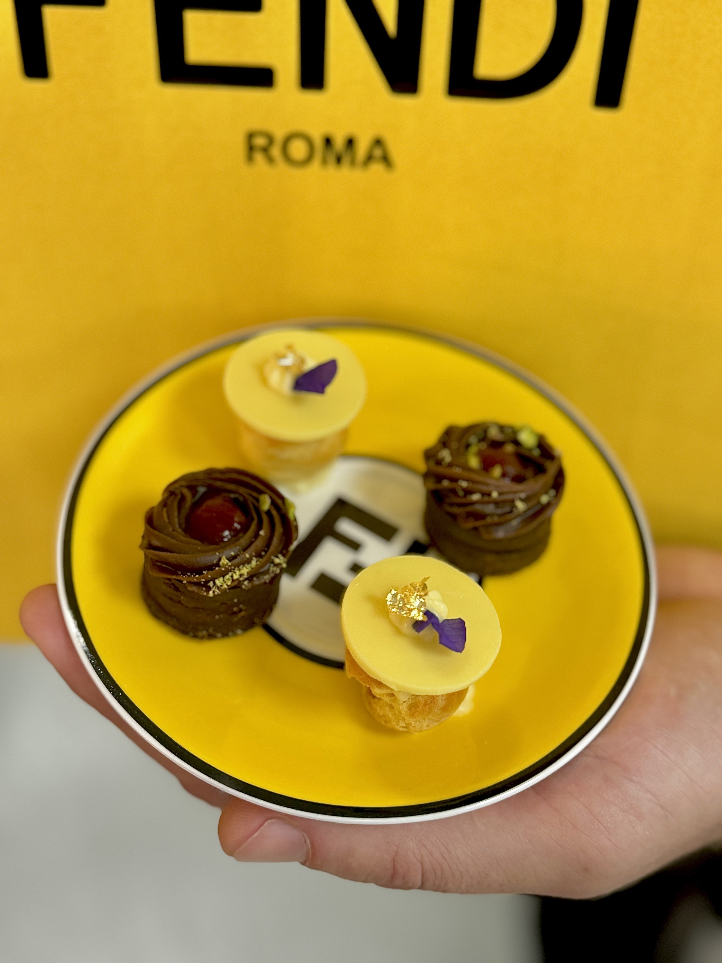Four small desserts on a yellow plate with a black and white logo, held by a person's hand. Two are chocolate with a cherry on top, and two are yellow with purple and gold decoration.