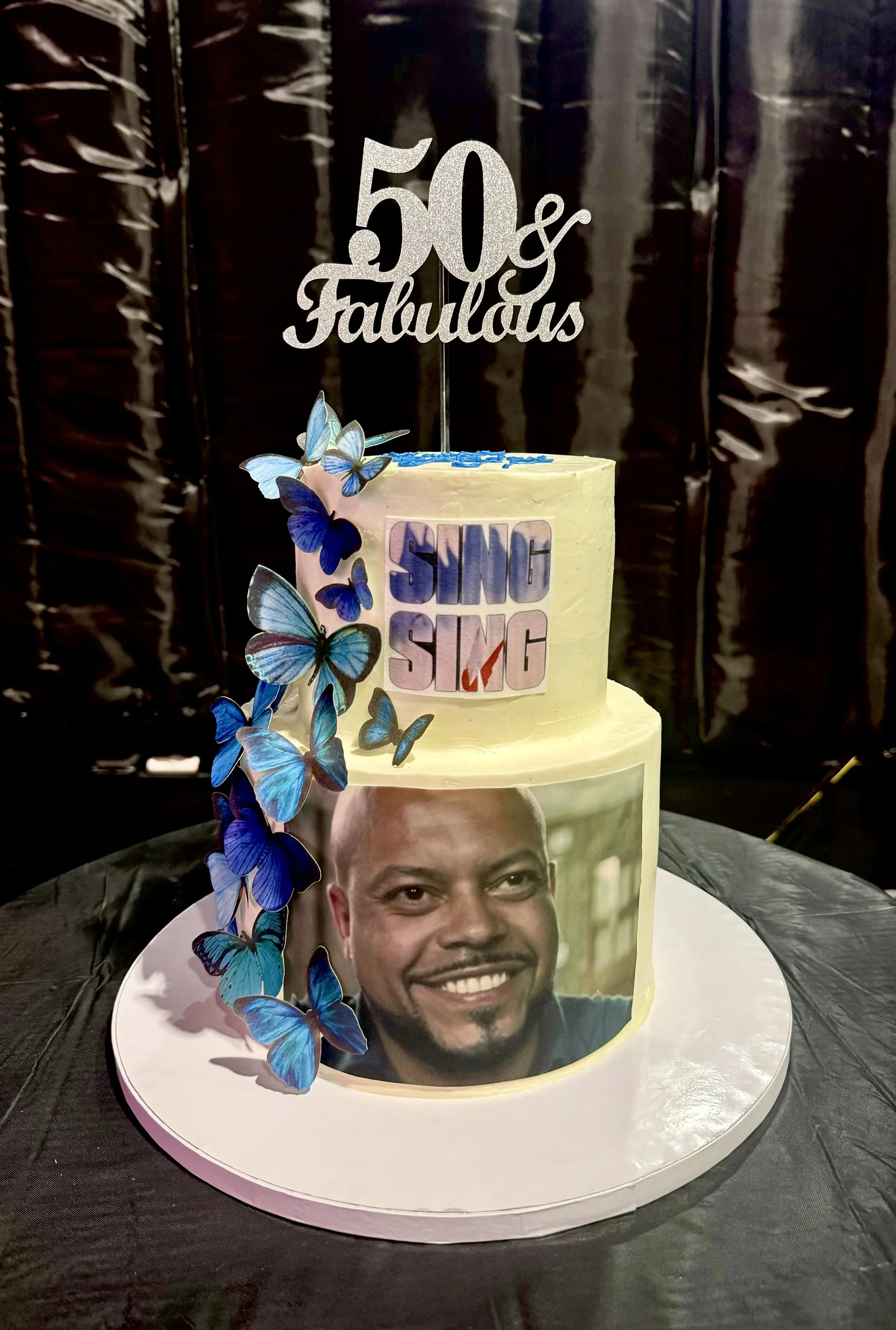 A two-tiered birthday cake with a photo of a smiling man on the bottom tier, decorated with blue butterflies on the side. The top tier has a 'Sing Sing' logo and a glittery cake topper that reads '50 & Fabulous.'