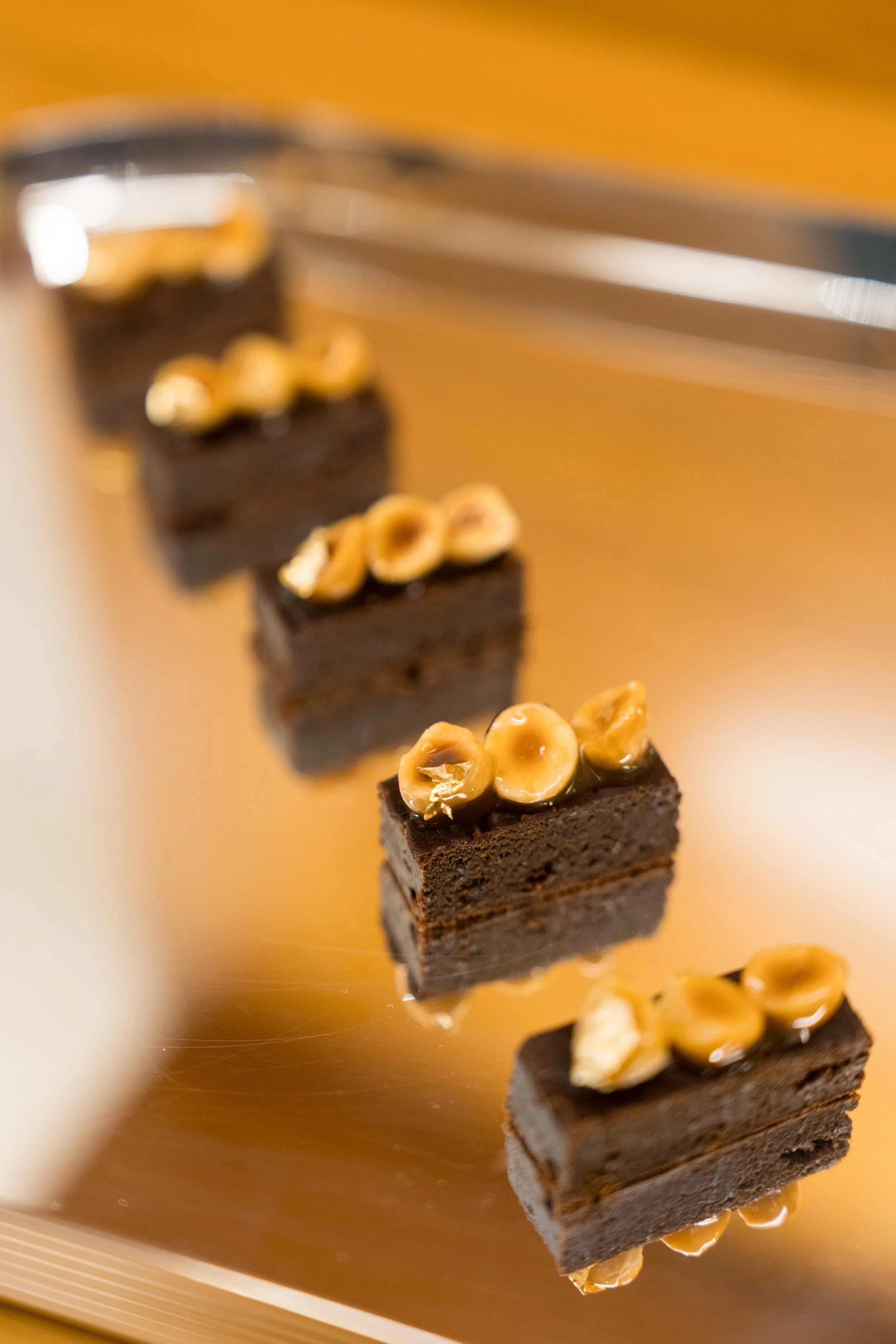 Close-up of four small chocolate cake squares topped with caramelized hazelnuts, arranged on a reflective surface.