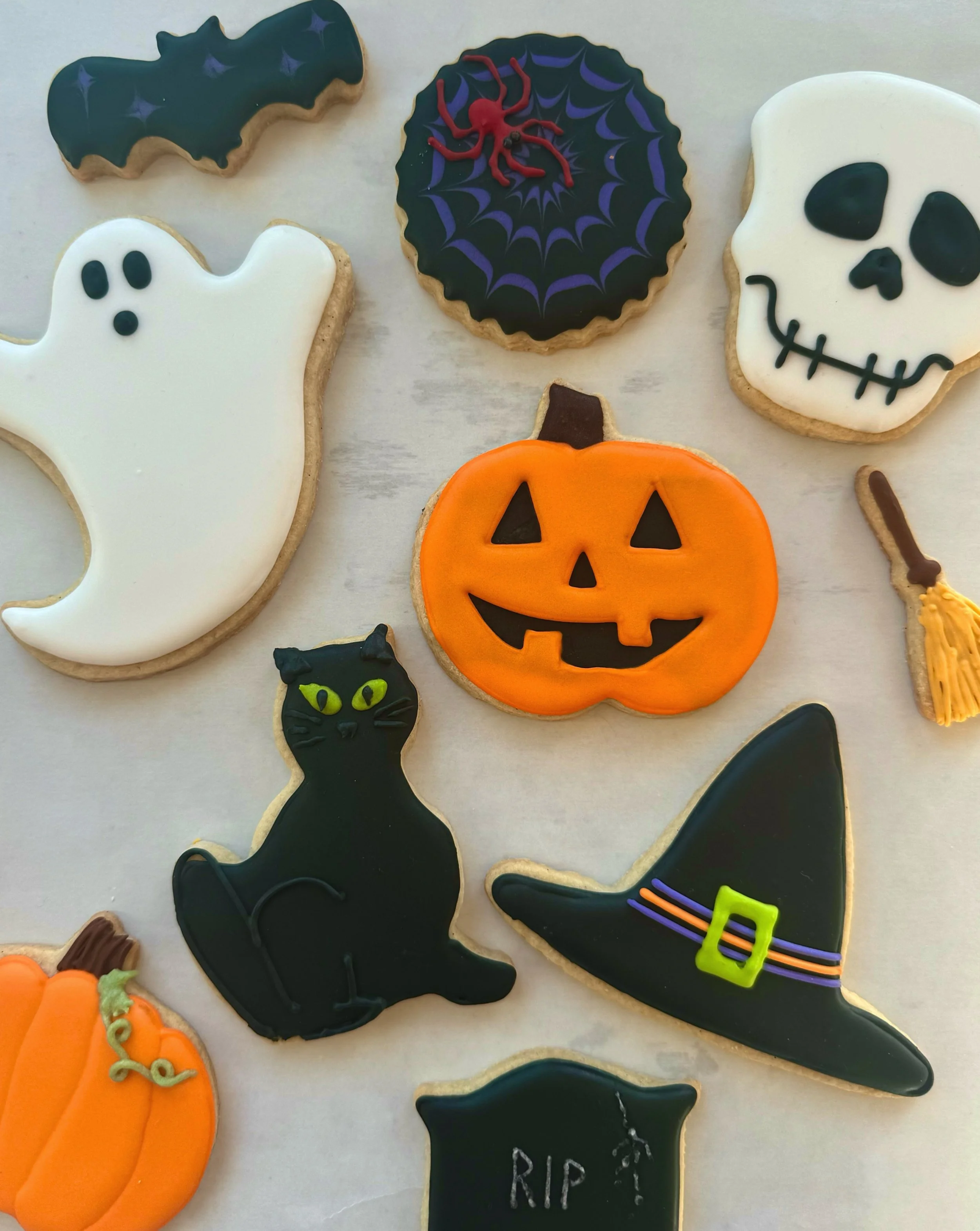 Assorted Halloween-themed decorated cookies, including a ghost, a black cat, a pumpkin, a skull, a bat, a spider on a web, a witch's hat, a tombstone, a broomstick, and a round cookie with spider web design.