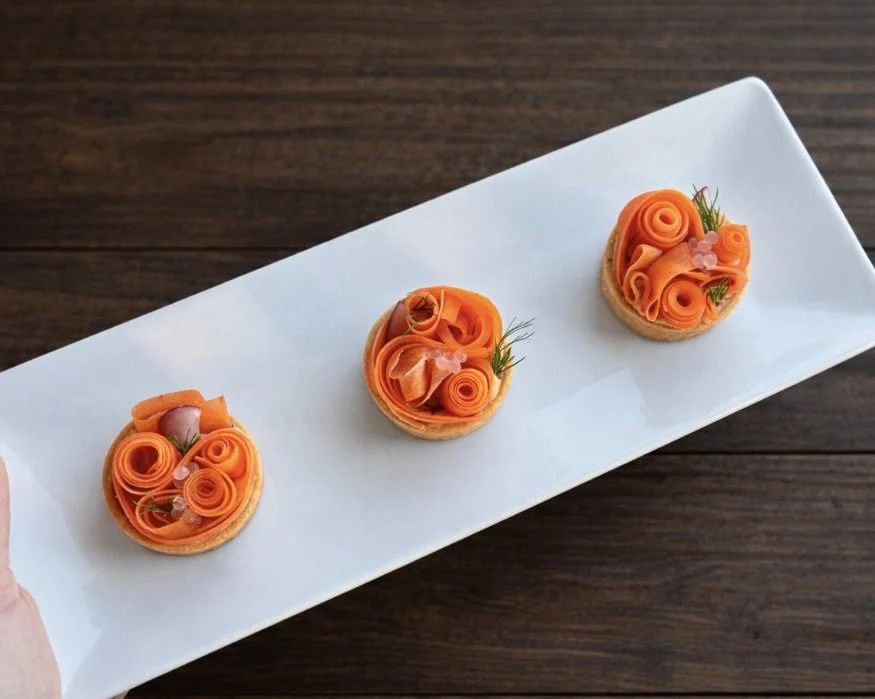 Three small tartlets topped with thinly sliced smoked salmon, rolled and curled to resemble flowers, garnished with herbs and small edible decorations, on a white rectangular plate.