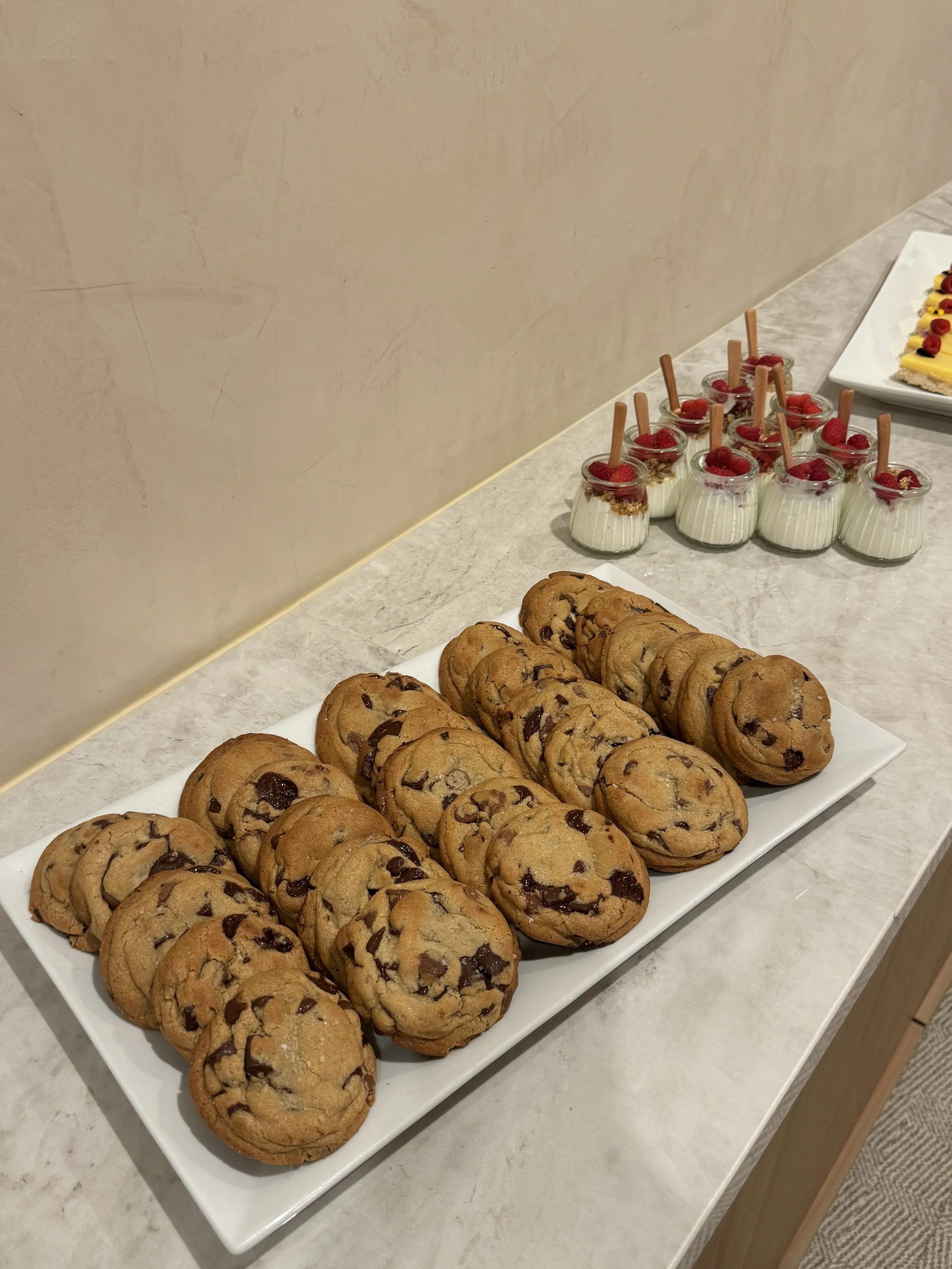 A white rectangular platter with chocolate chip cookies on a marble countertop. In the background, there are small glass cups with panna cotta topped with raspberries and a dessert with berries and cream on a white plate.