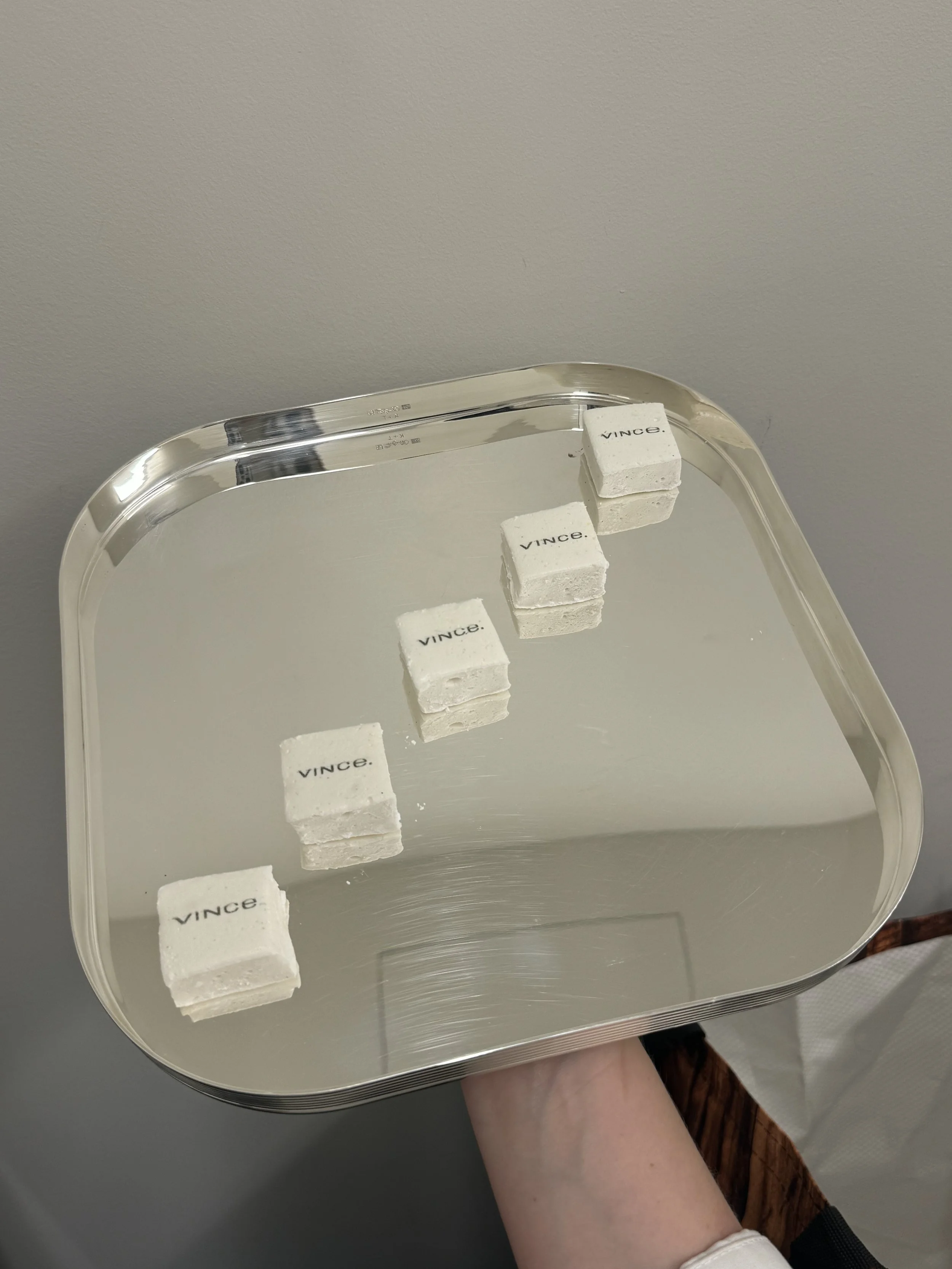 Four small white cubes with the word "VINCE" printed on top are arranged in a line on a silver tray, reflected in the tray's surface.