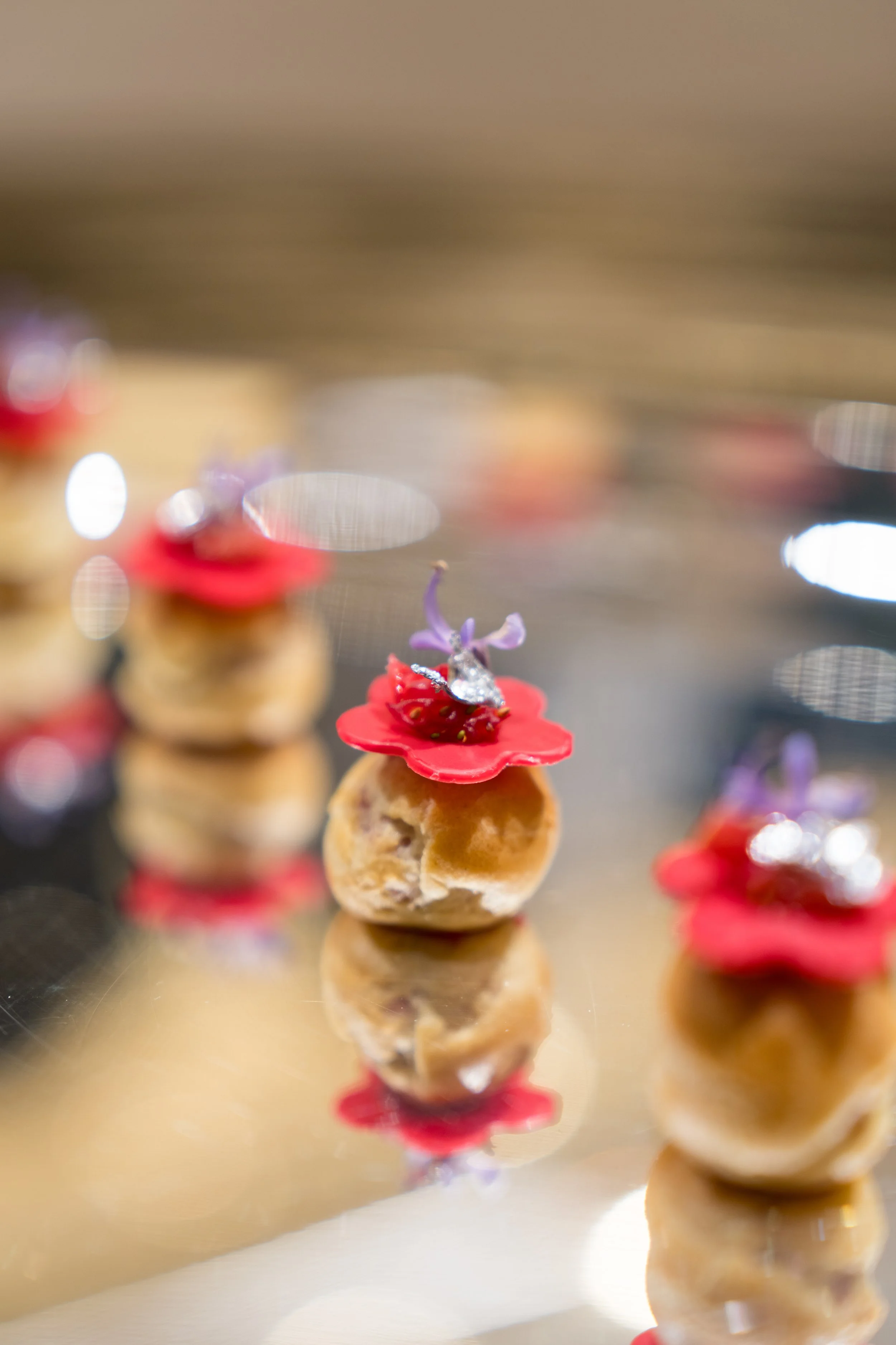 Close-up of small dessert pastries topped with purple flowers, red edible accents, and a silver decorative element, reflected on a shiny surface.