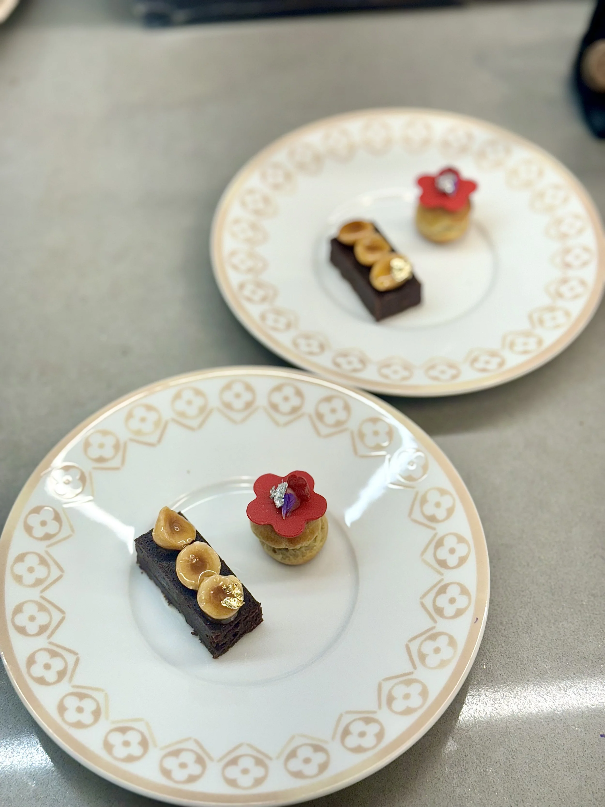 Two decorative plates each with two small desserts, one chocolate cake with gold flecks and almonds, and a tiny pastry with a red flower-shaped topping and a small silver decoration, on a gray surface.