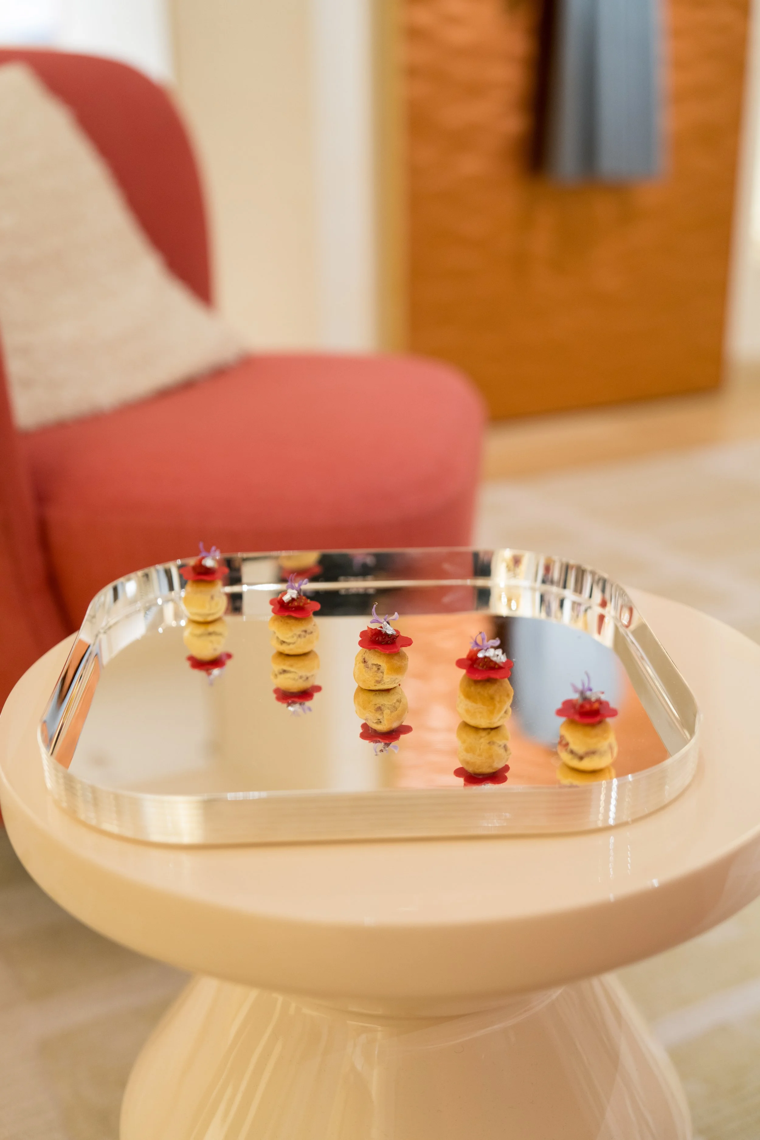 Six small, round desserts topped with edible flowers and red sauce, arranged on a reflective metal tray on a round white table.