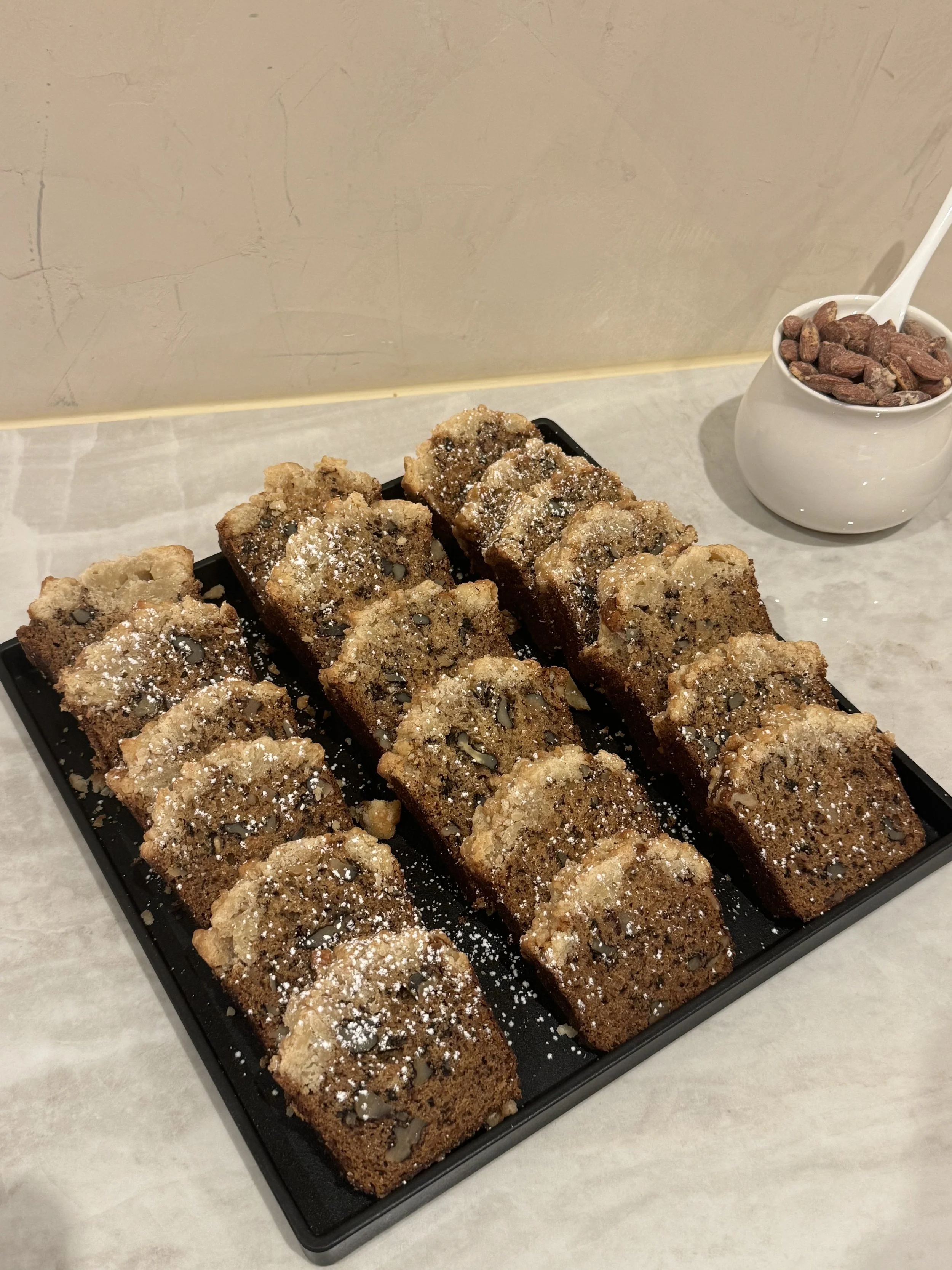 A black tray of sliced chocolate chip banana bread topped with powdered sugar on a table, with a white bowl of cocoa beans if off to the side.