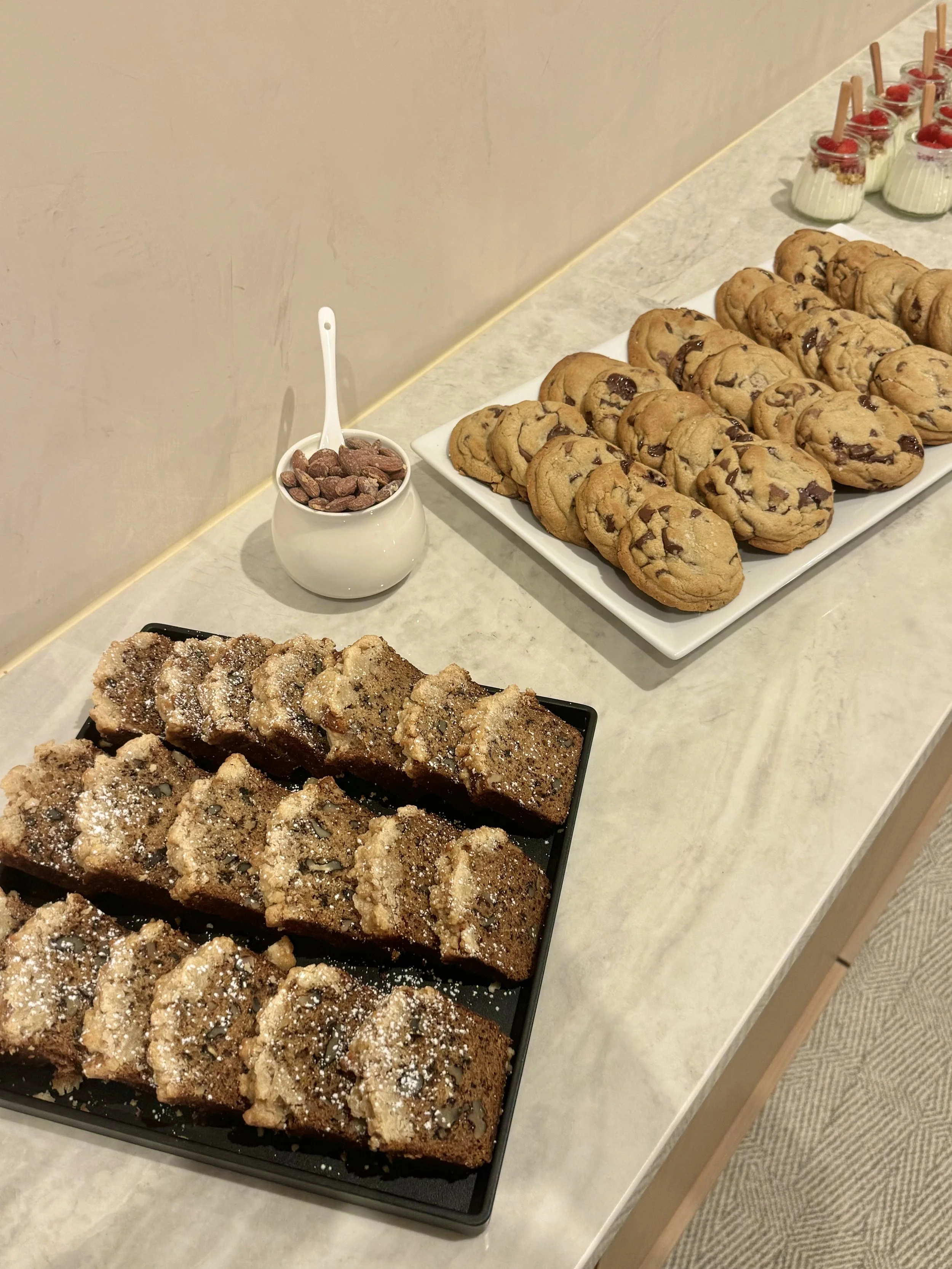 Assorted baked goods, including chocolate chip cookies, crumb-topped coffee cakes, and small jars of pudding topped with cherries, arranged on a white counter.