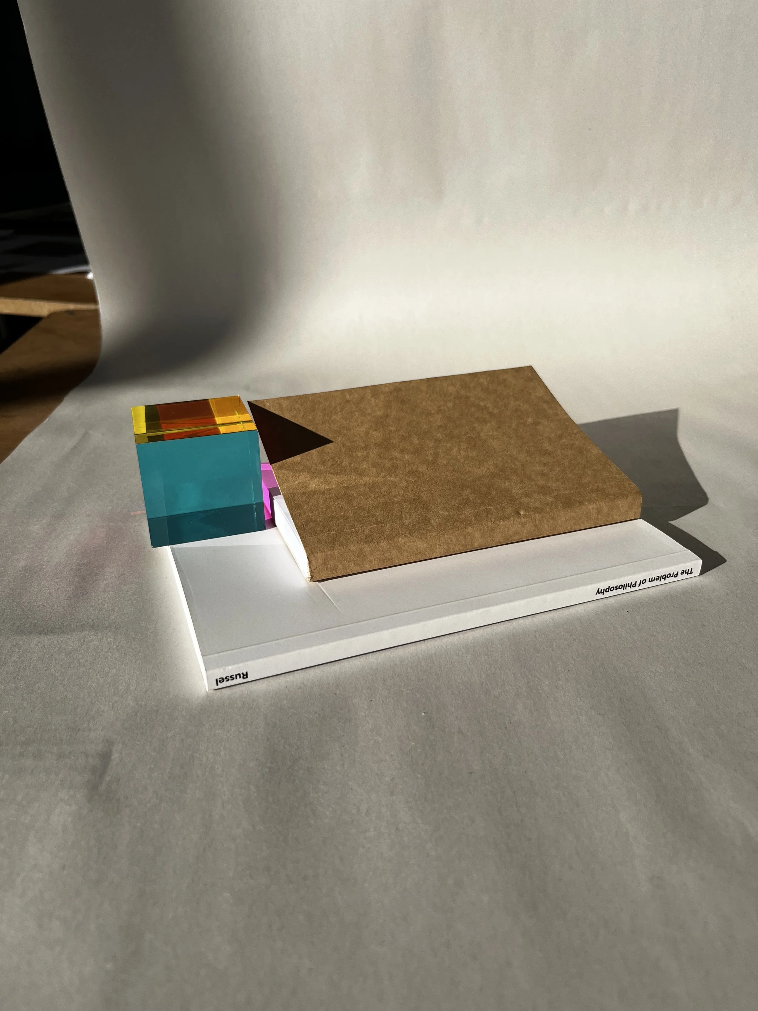 A white board with a small, colorful, geometric paper sculpture and a brown paper square resting on it, with shadows cast by the objects.