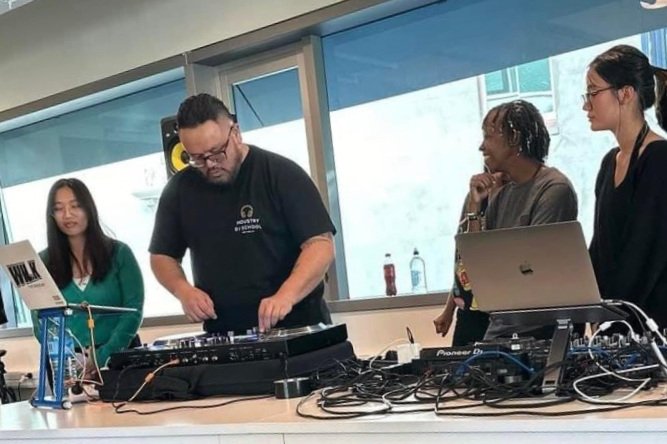 A DJ performs in front of a laptop and mixing equipment while four women watch, inside a room with large windows.