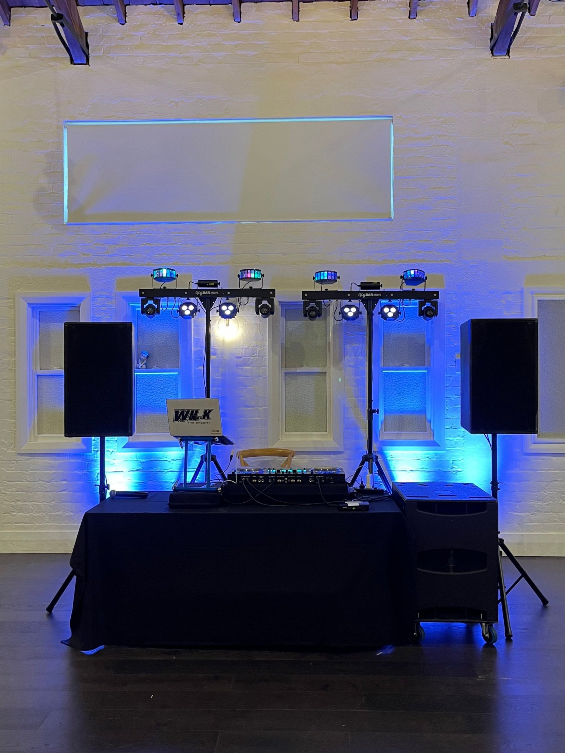 DJ setup with speakers, lighting equipment, and a table with a laptop in a room with white brick walls and blue lighting.