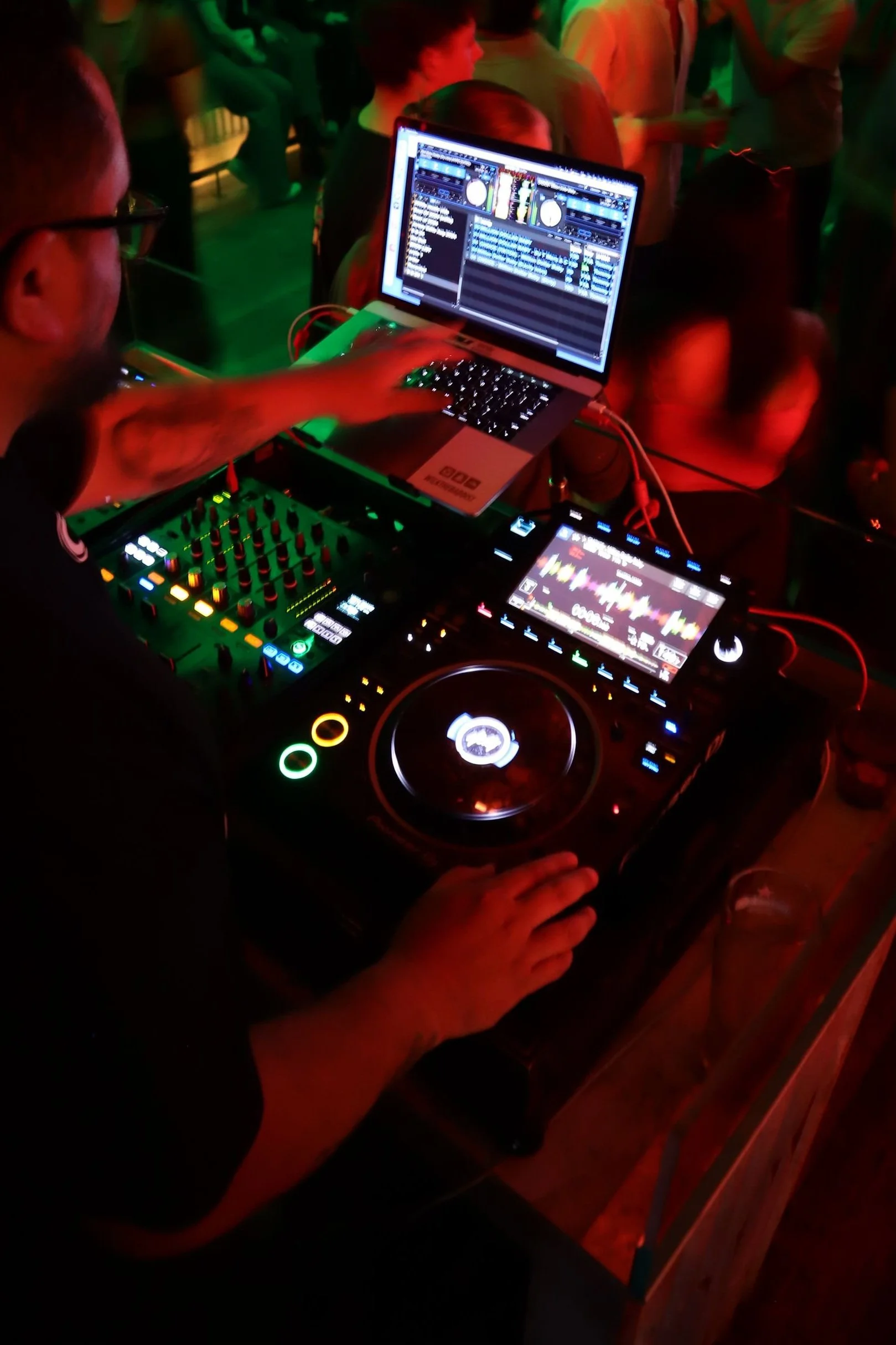 DJ mixing music at a nightclub with a laptop, DJ controller, and digital audio equipment on a table, surrounded by a crowd of people.