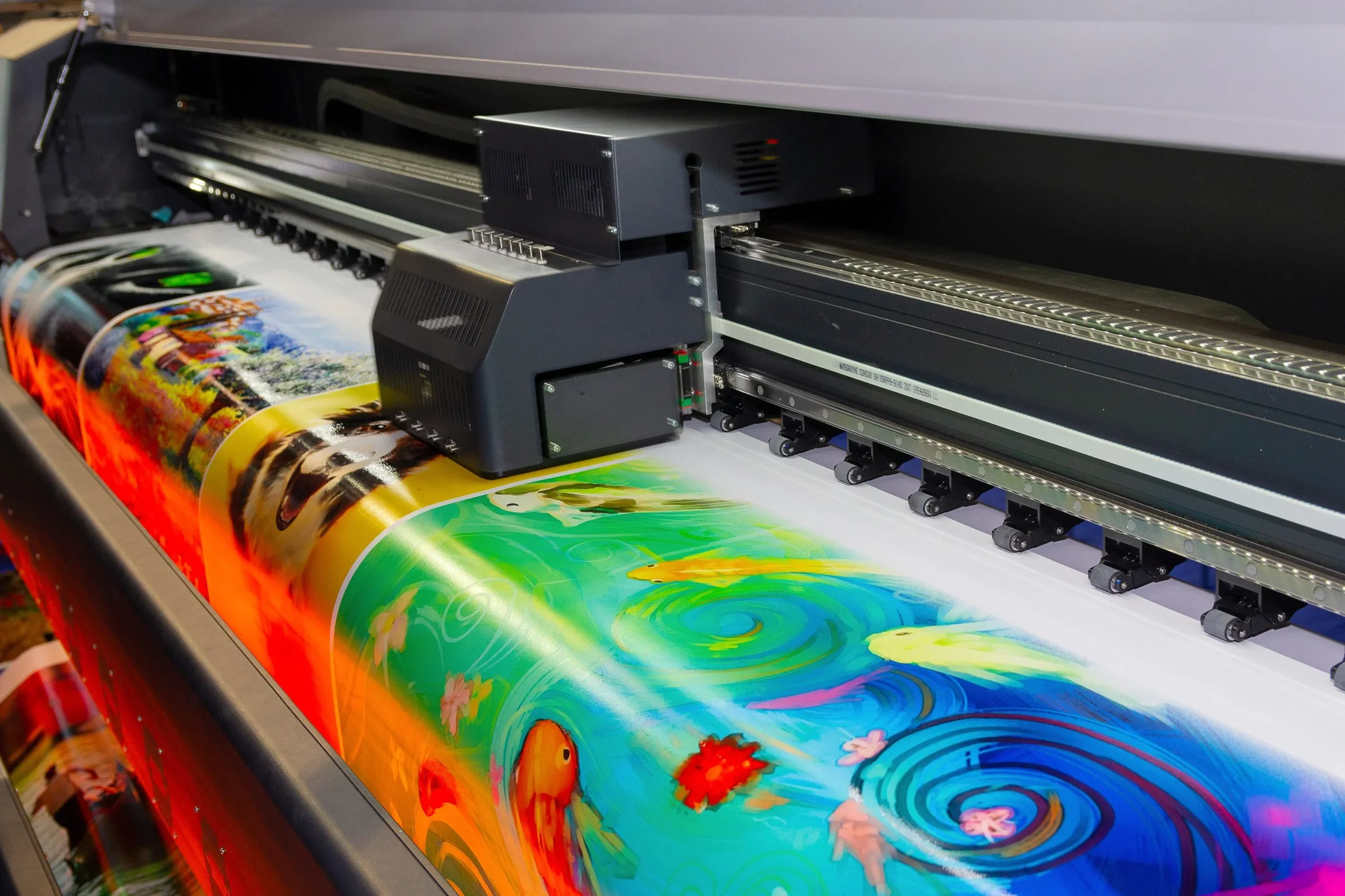 A large-format printer printing a colorful, vibrant image of a pond with fish, flowers, and swirling water patterns on a long roll of paper.