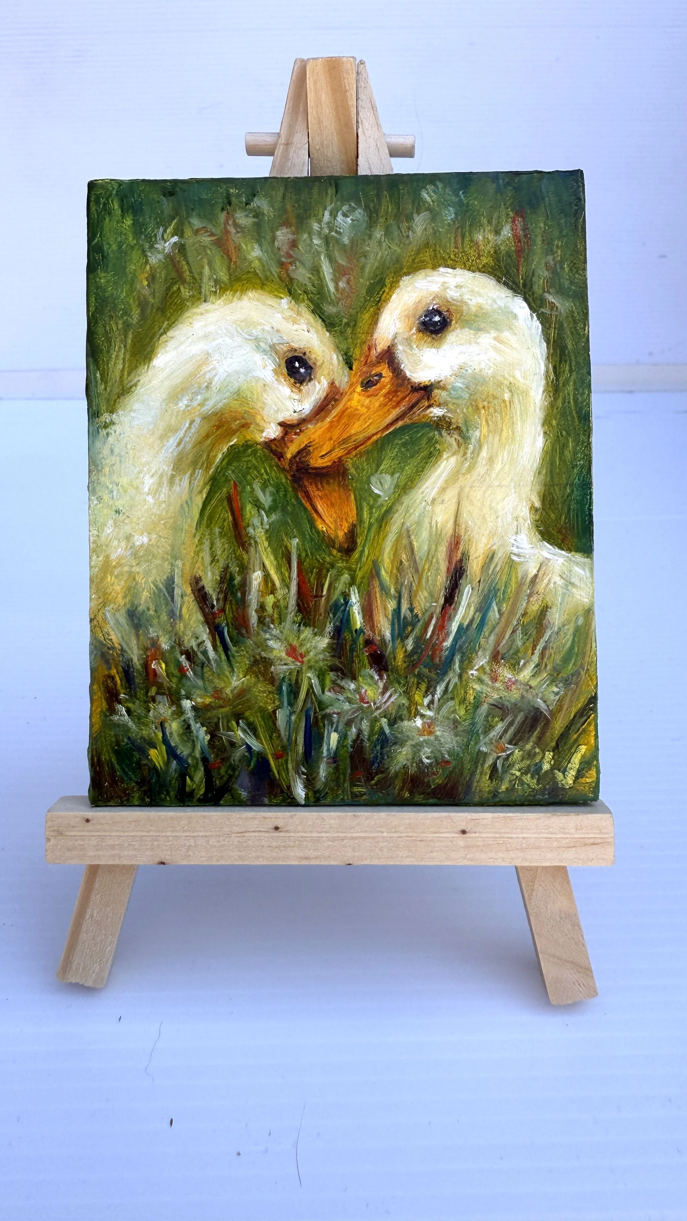 Mini oil painting of two white geese touching beaks in tall grass, on a wooden easel.