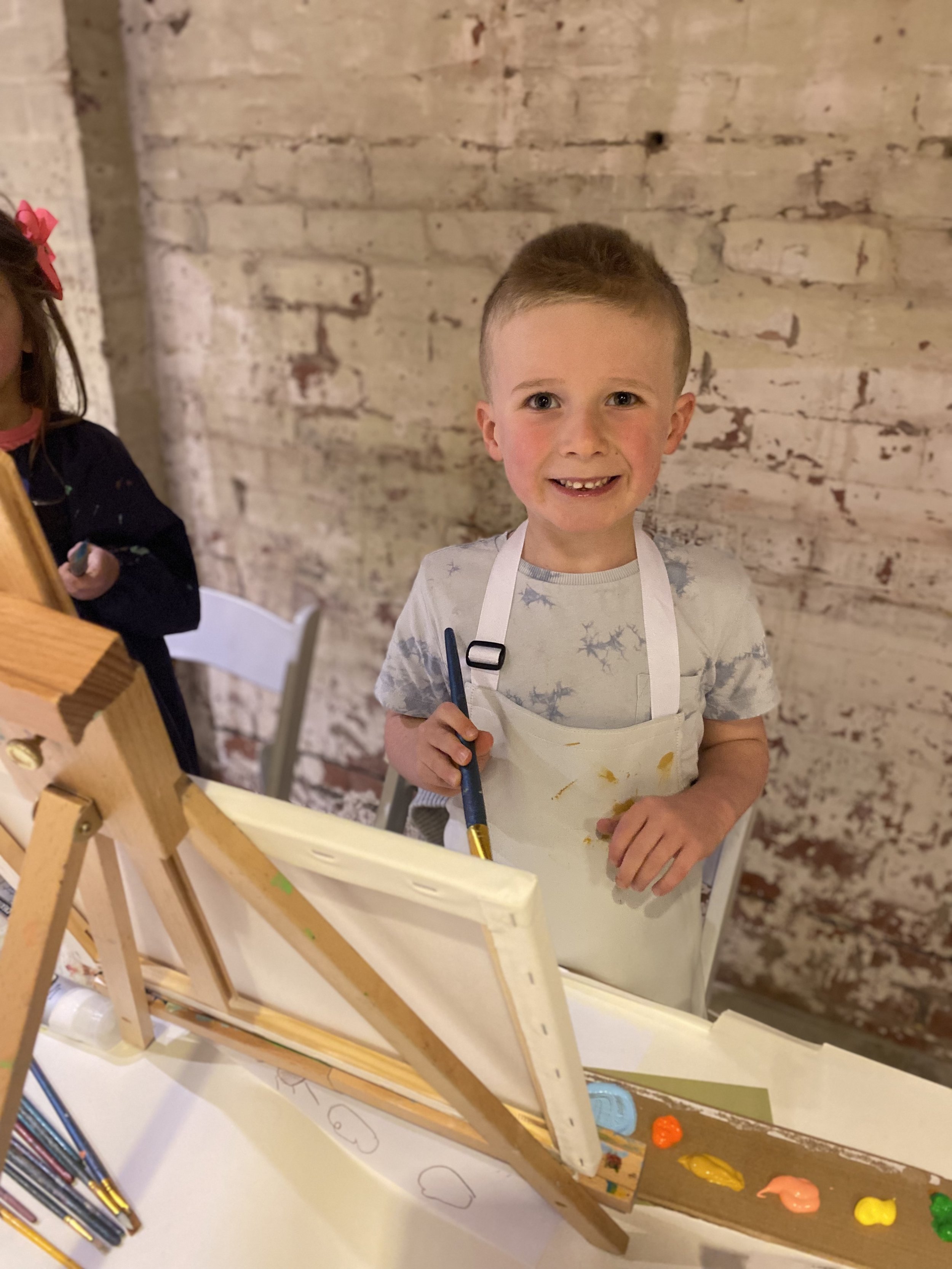 A young boy with short hair, wearing a paint-stained apron, stands smiling at an easel with paints and brushes, in front of a brick wall.