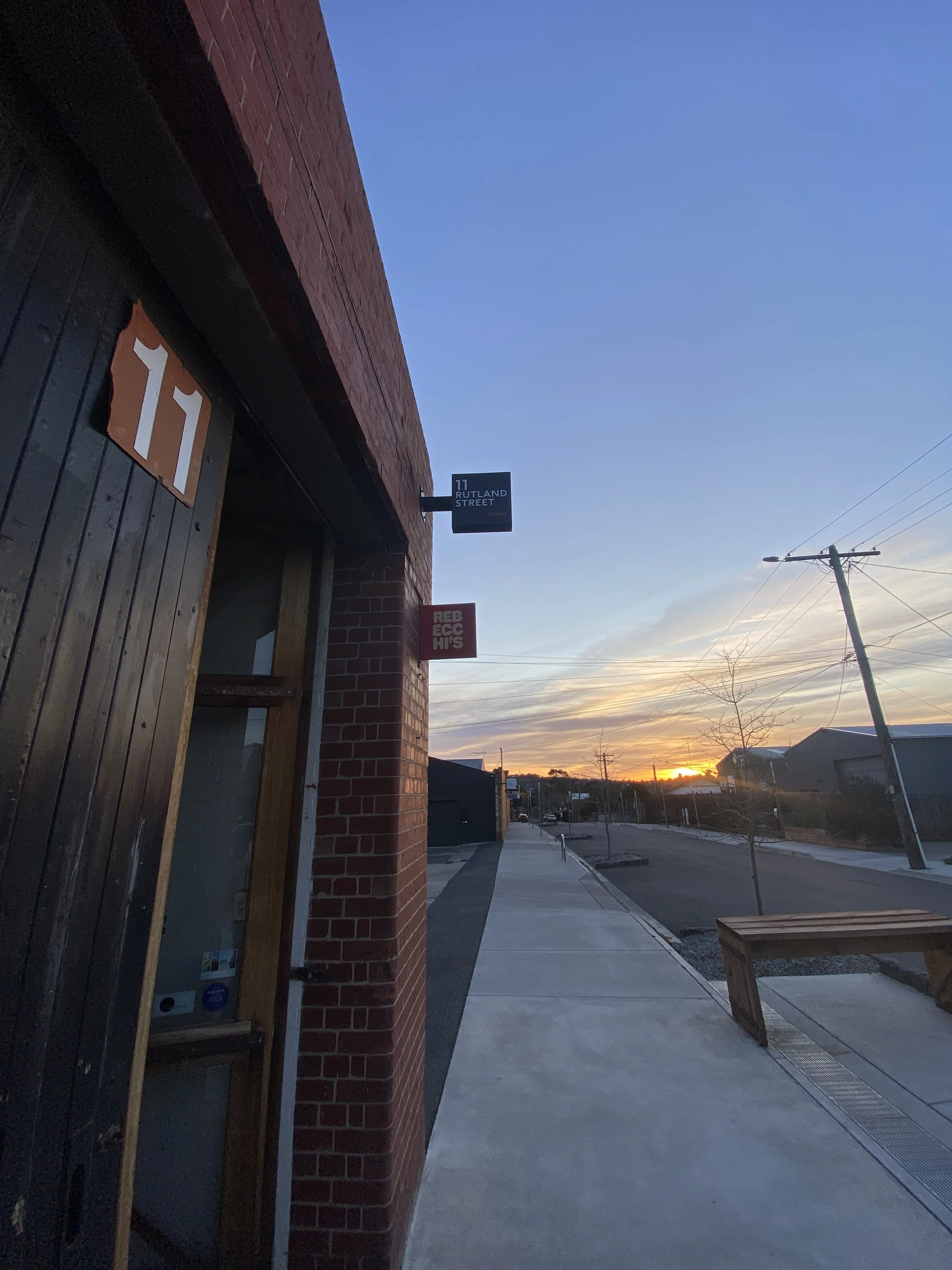Street view at sunset with a brick building on the left, indicating the address 11, and a sign for Rutland Street. The sidewalk is empty, with some small trees and a bench on the right, and power lines stretching across the sky.