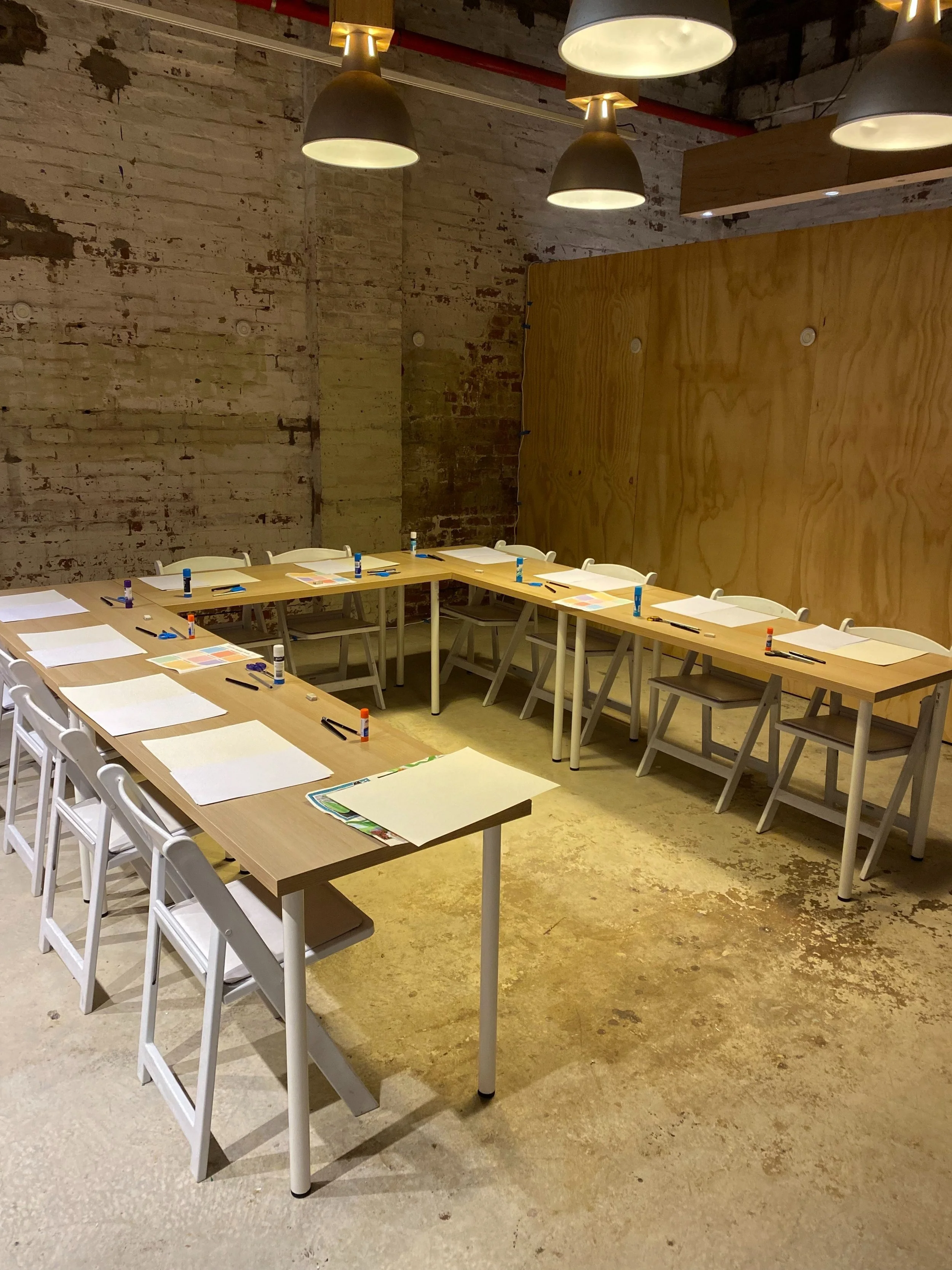 A conference room or classroom setup with tables arranged in a U-shape, each with paper, markers, and glue sticks. There are white chairs around the tables, industrial-style hanging lights overhead, and a mix of exposed brick and wooden wall surfaces.