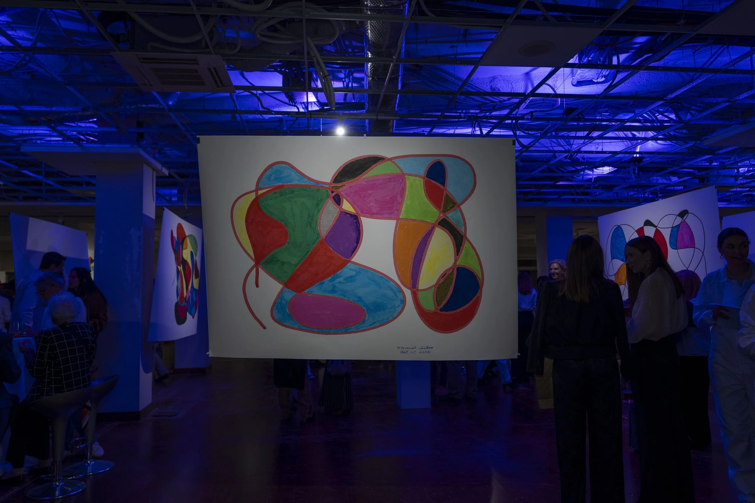Obra de arte abstracta con formas coloridas y líneas curvas, exhibida en un ambiente con iluminación azul y personas observando en un espacio de galería o exposición.