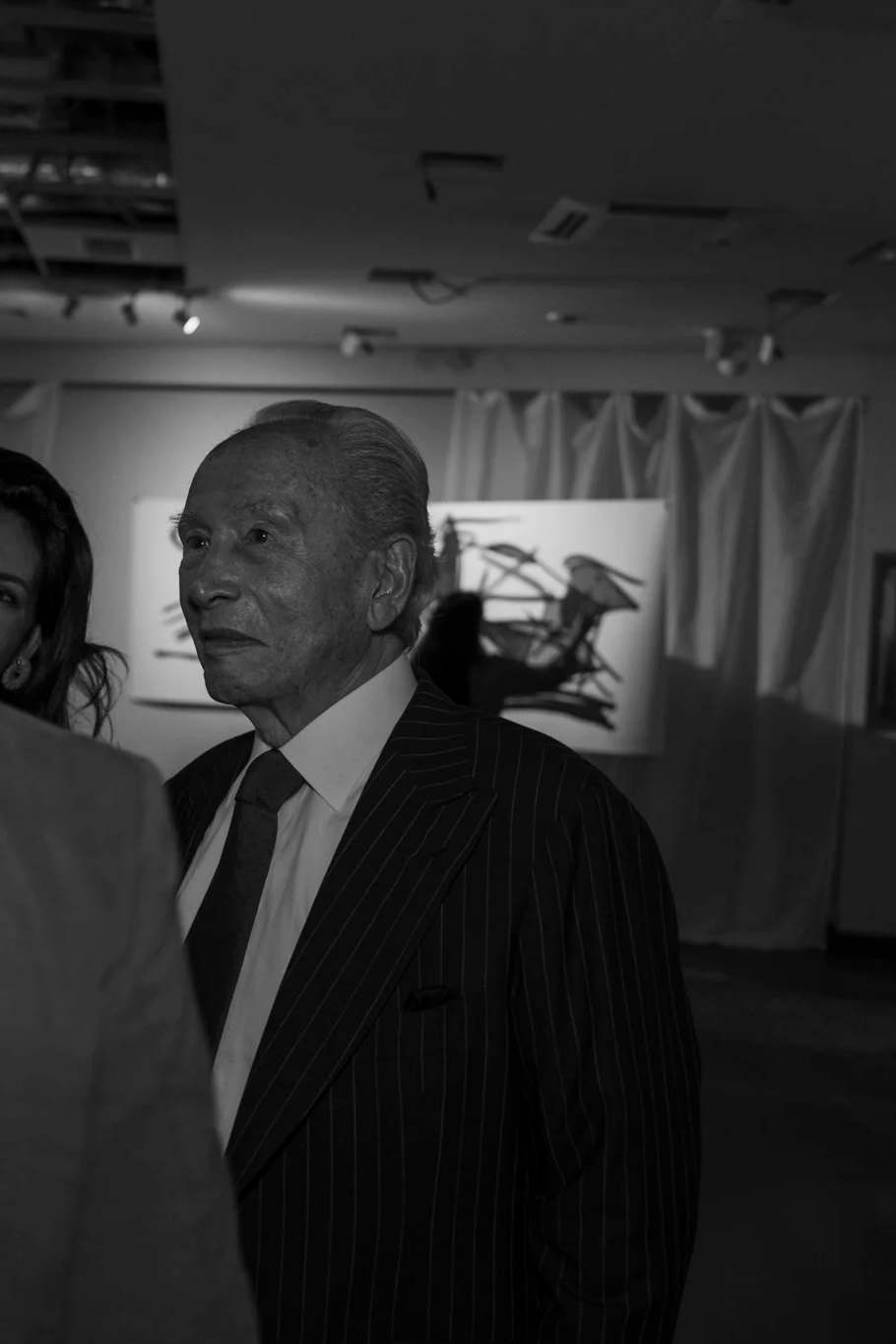 Hombre mayor con cabello gris peinado hacia atrás, con traje de rayas y corbata, en un ambiente de exposición de arte, en un espacio con cortinas blancas y una obra abstracta en el fondo.