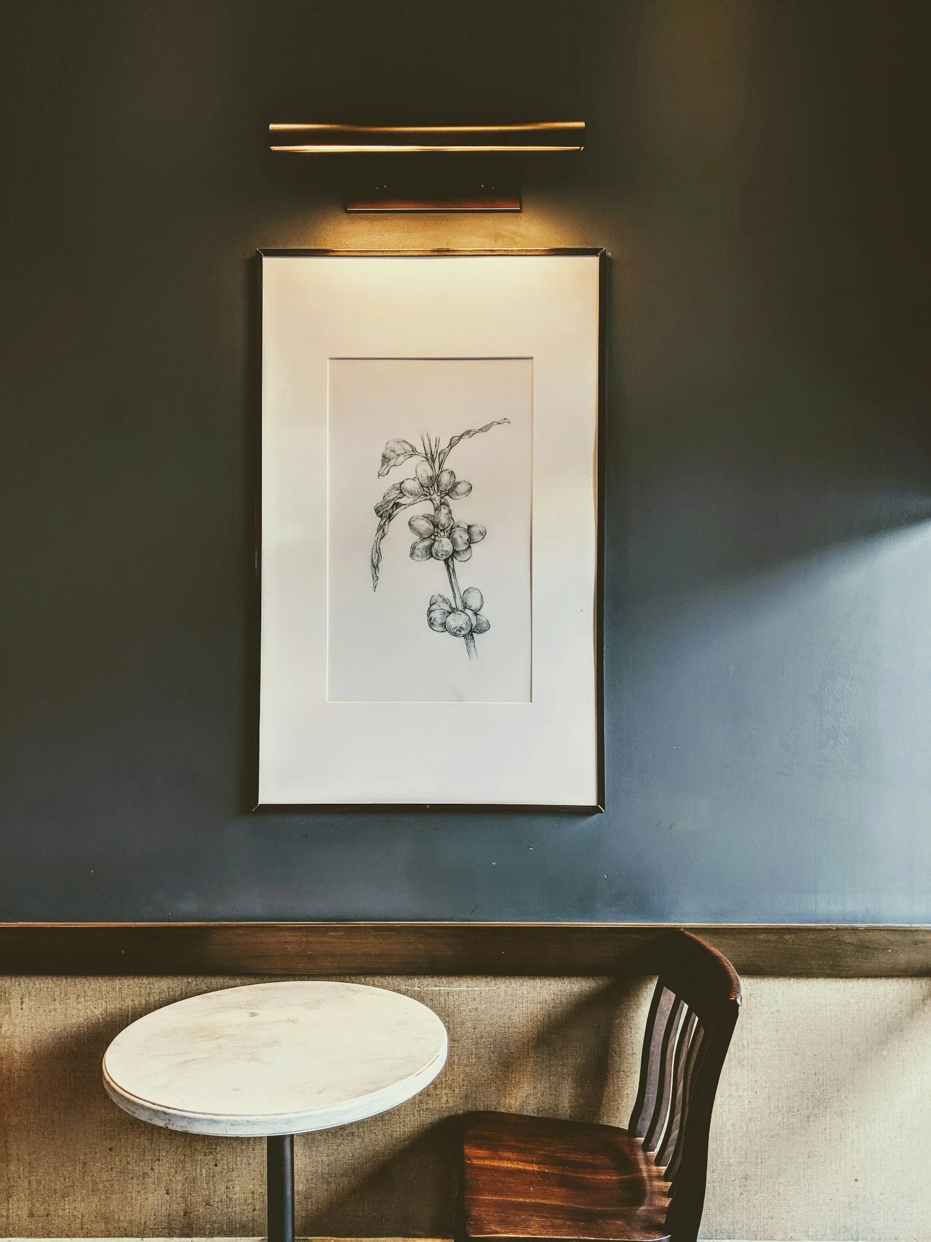 A framed black and white botanical sketch of a branch with berries hangs on a dark gray wall. Below the art, a small round table with a light-colored top and a dark metal base sits next to a wooden chair with a curved backrest.
