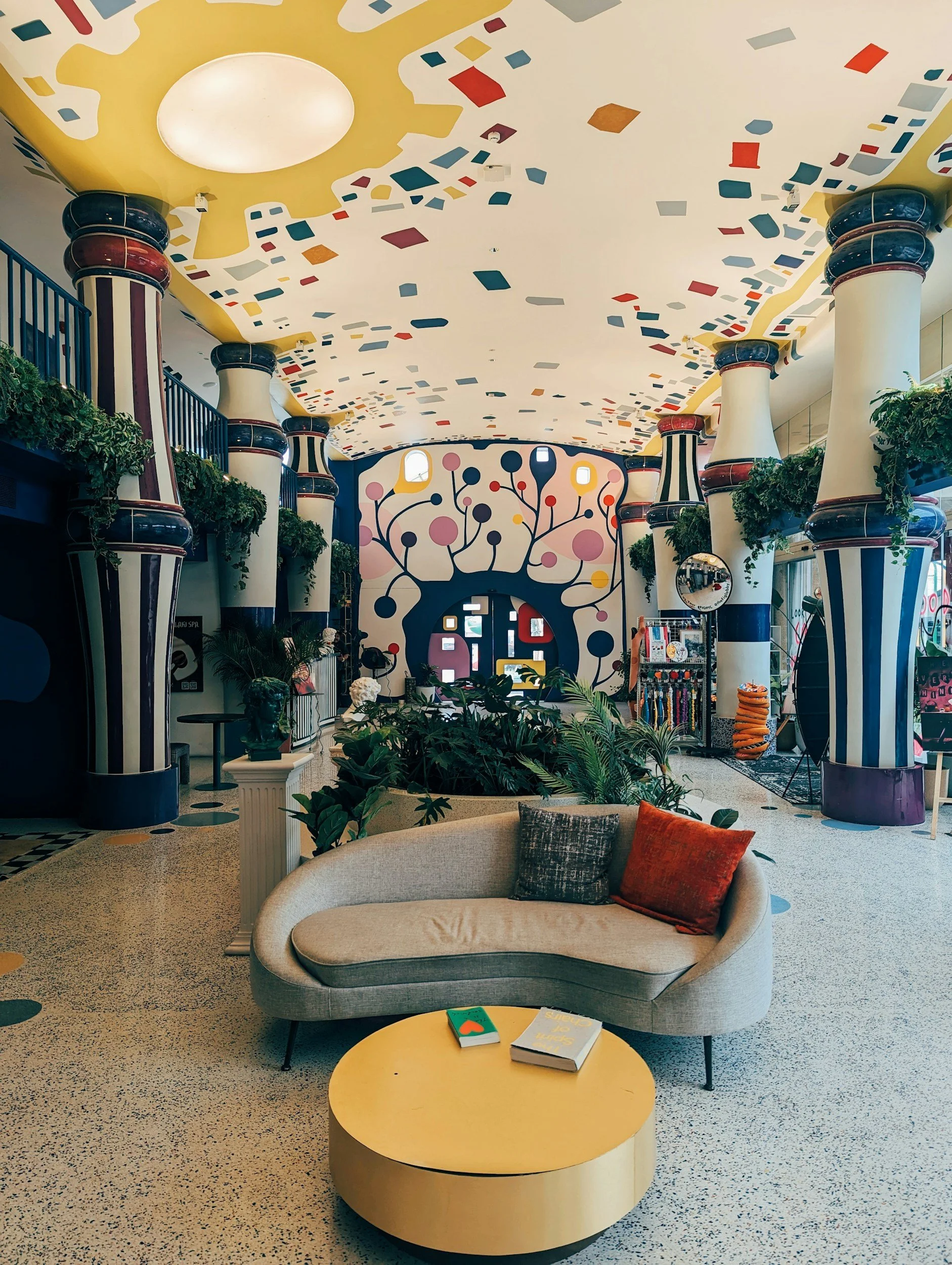 Interior of a colorful, modern lounge with curved furniture, lush plants, and vibrant wall art featuring a tree with round pink and dark blue leaves.