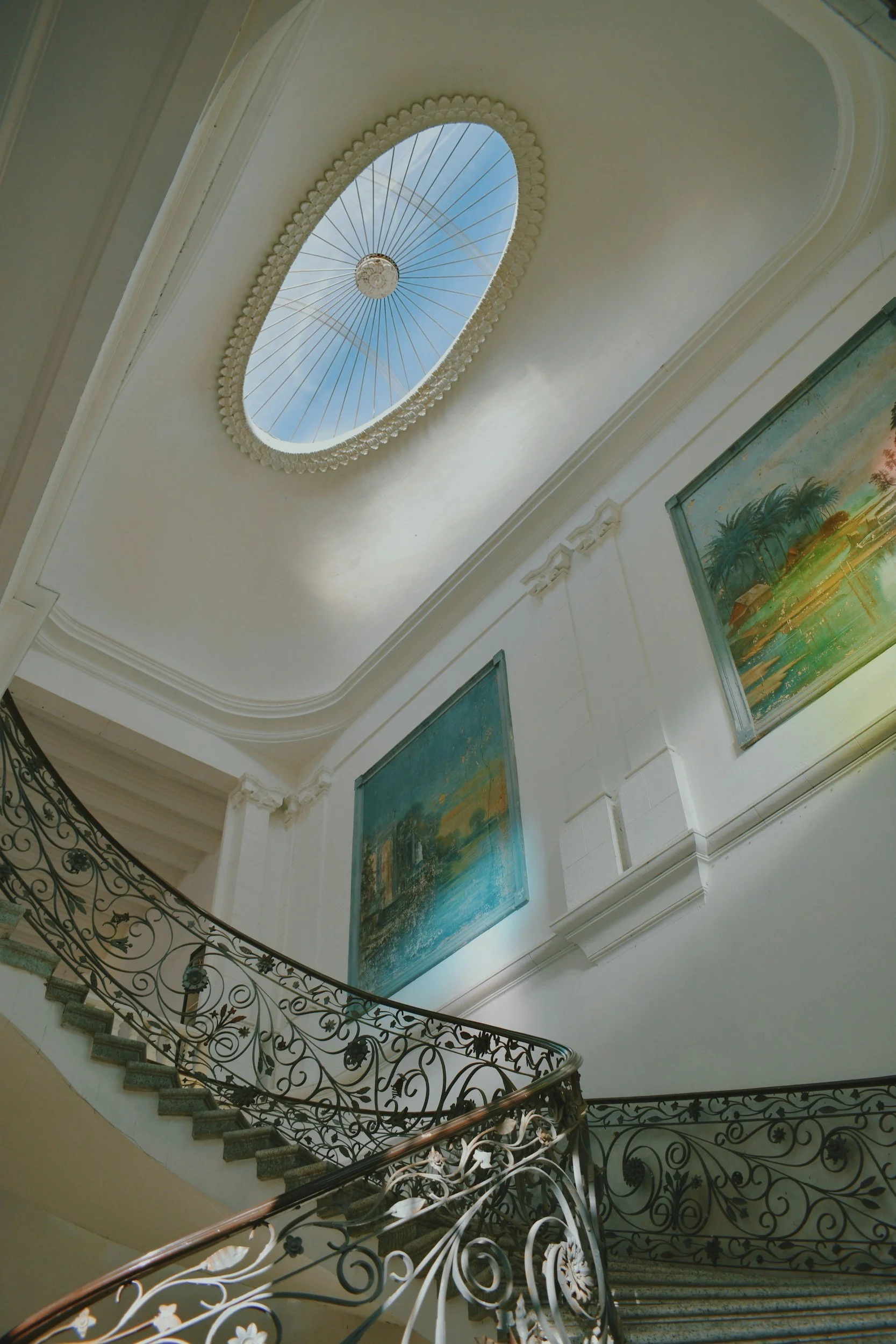Interior view of a staircase with ornate wrought iron railing, leading up towards a white ceiling with a skylight, and two colorful landscape paintings on the wall.