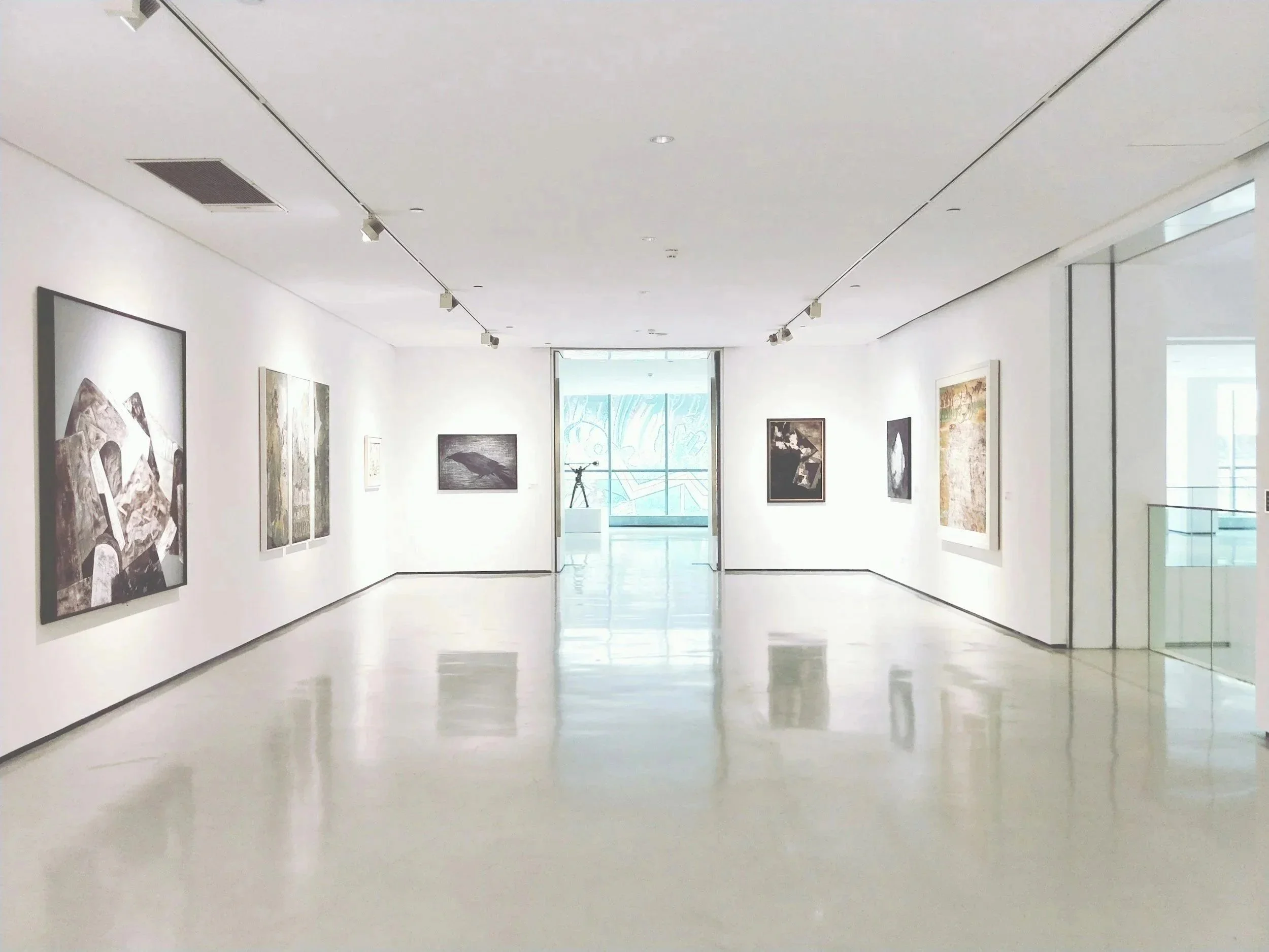 Empty art gallery with framed paintings on white walls, a sculpture on a pedestal near the back wall, and large windows allowing natural light.