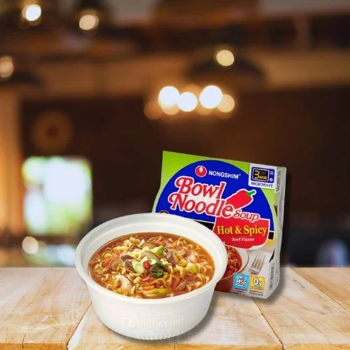 A bowl of hot spicy beef-flavored noodle soup with sliced green onions on top, placed on a wooden surface next to a package of NongShim Bowl Noodle Soup.