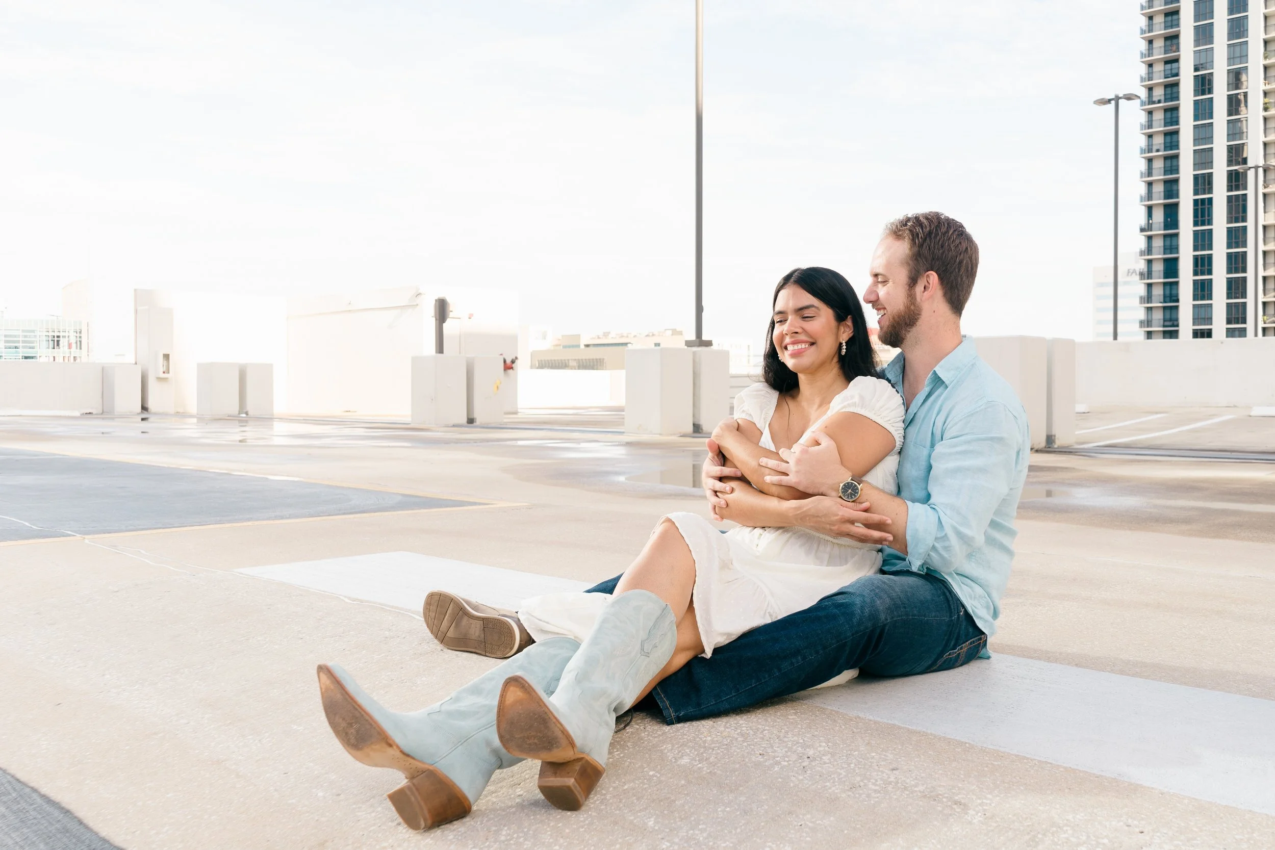 A happy couple sitting on a rooftop parking lot, embracing and smiling with city buildings in the background during daytime.