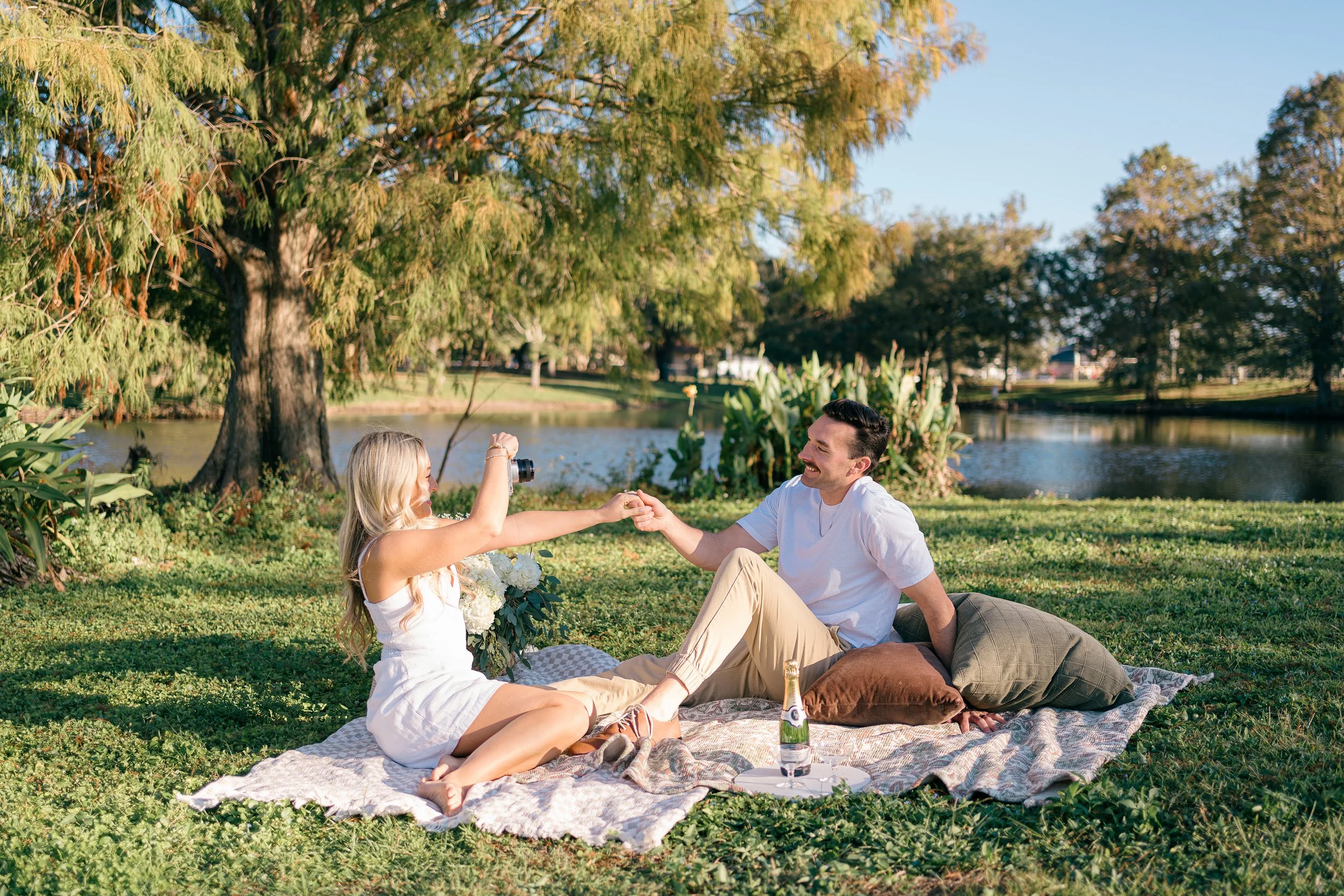 A couple having a picnic by a lake on a sunny day; woman taking a photo of man, who is smiling and holding her hand, sitting on a blanket with pillows, a bottle of champagne, and flowers.