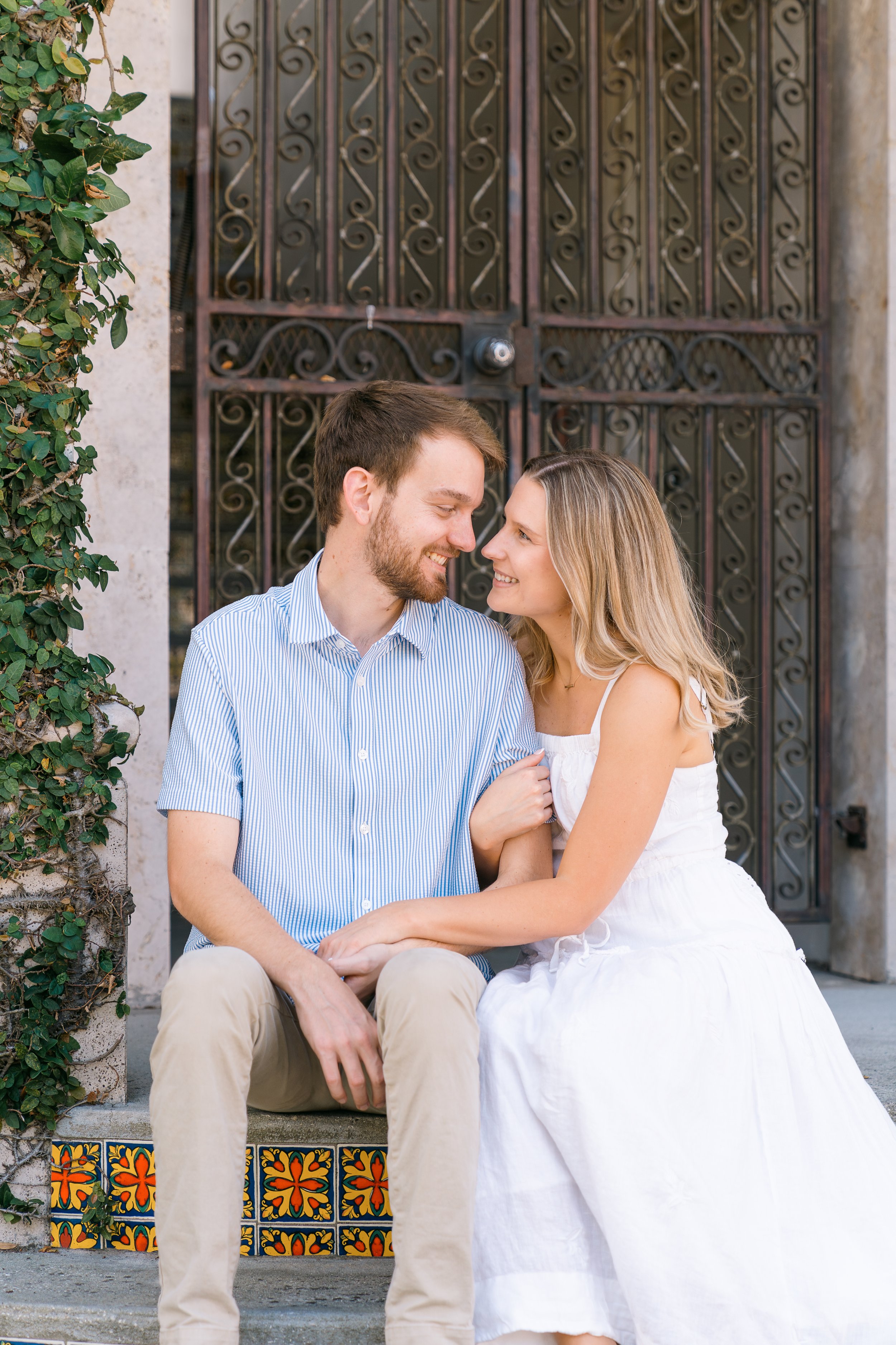 A young couple sitting on steps in front of a decorative iron gate, smiling at each other, with greenery on the side.