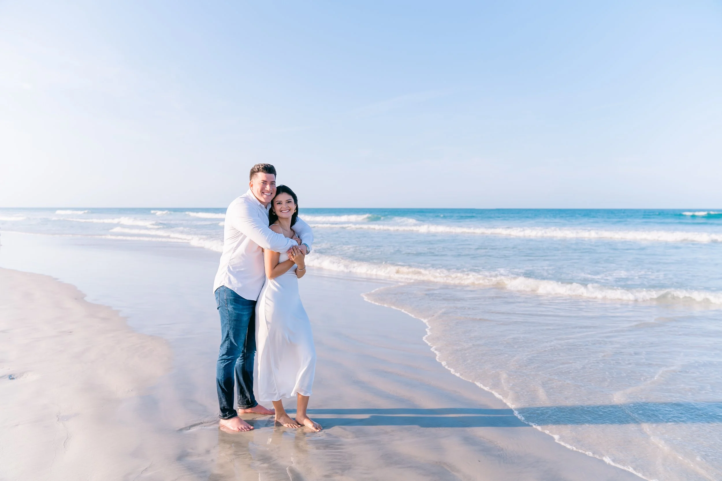 A couple hugging on a beach, with the ocean and blue sky in the background.