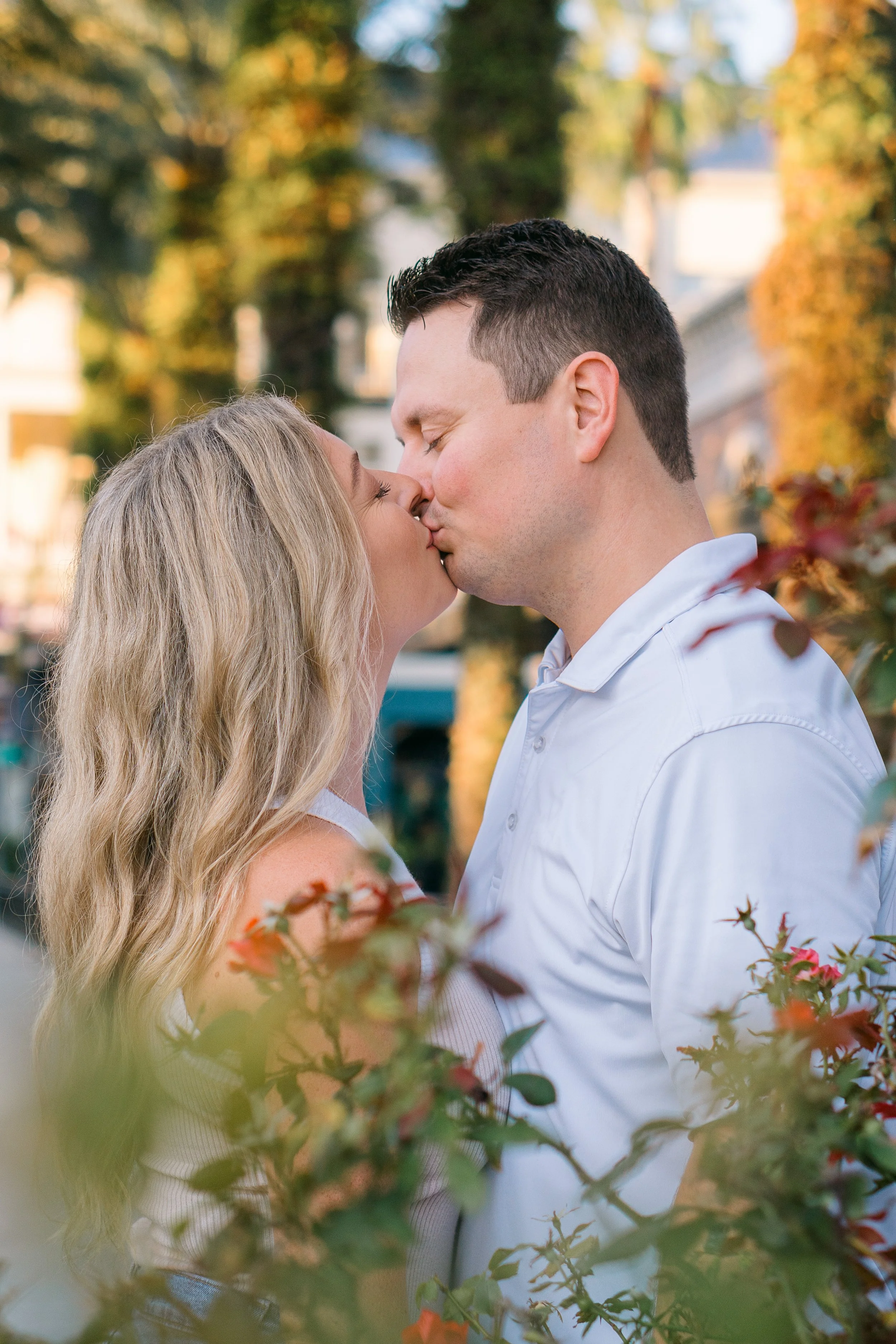 A couple kissing outdoors among trees with autumn foliage, with the woman having long blonde hair and the man wearing a white shirt.
